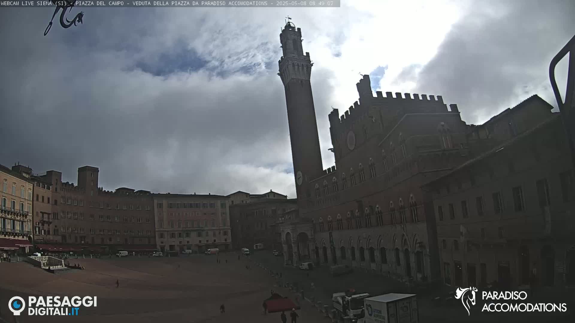 A large, mostly cloudy sky looms over a wide, open plaza in a city, where several historic buildings, including a tall tower and a large palace, stand.