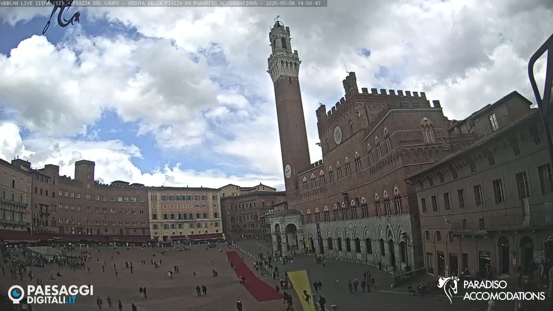 A partly cloudy sky overlooks a large plaza with several brick buildings, including a tall tower and a palace, and a red carpet extending across the center of the plaza.