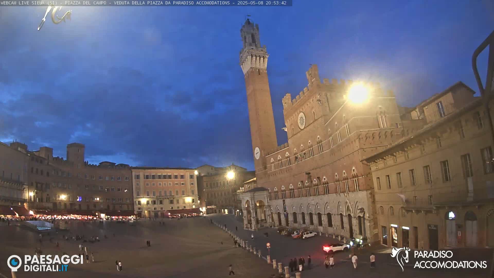 A large, mostly empty town square at twilight is surrounded by medieval-style buildings, including a tall brick tower and a palace.