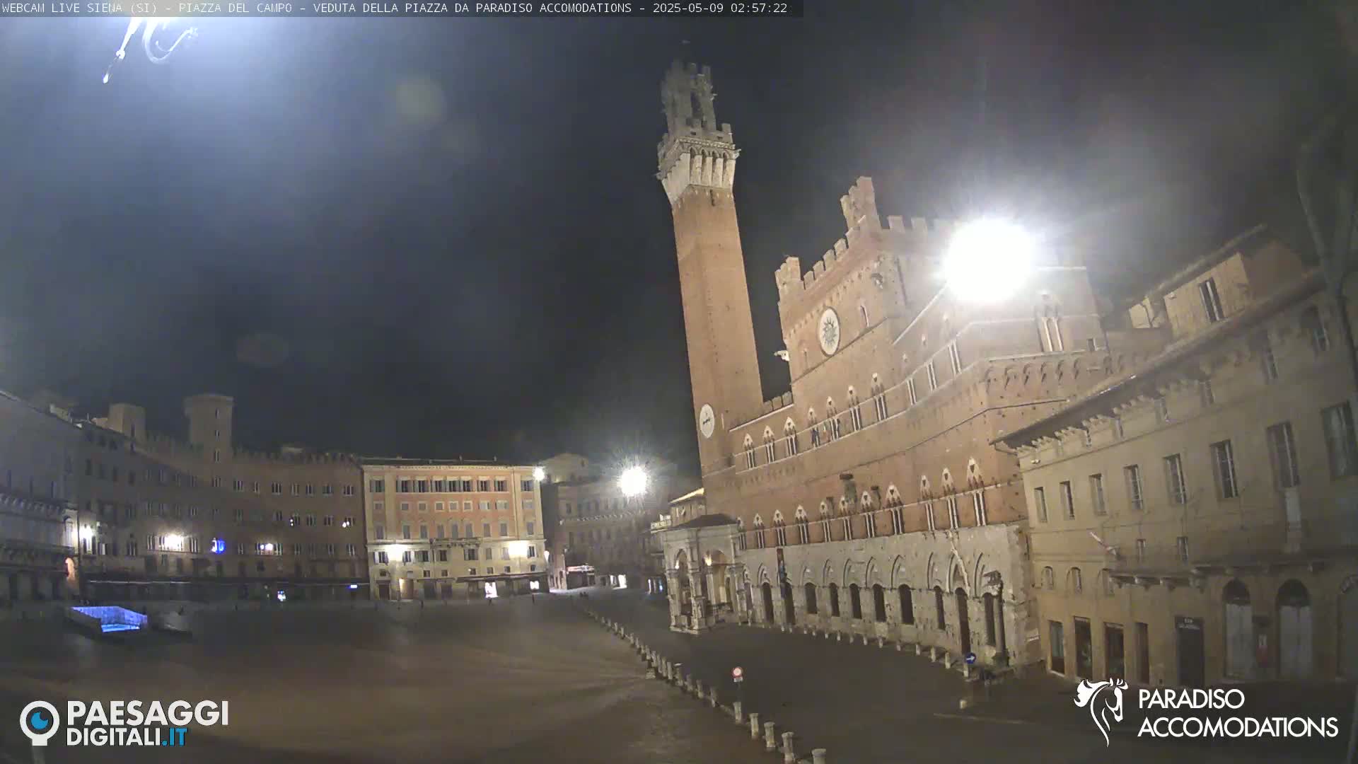 A large, empty plaza at night, illuminated by streetlights, is centered on a brick building with a tall tower.