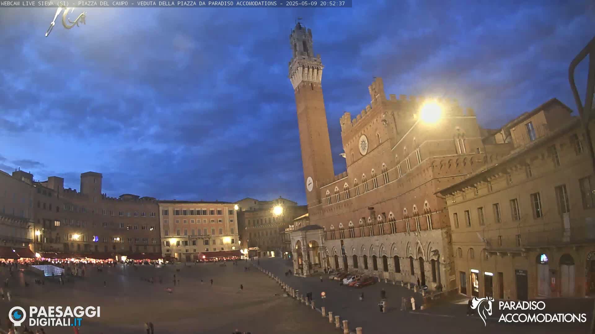 A large, mostly empty piazza in an Italian city at twilight, surrounded by historic brick buildings, one featuring a tall tower.