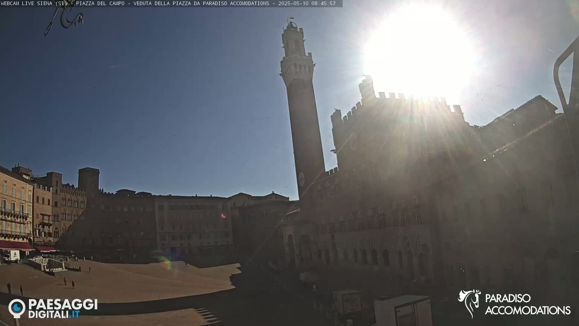 A sunny day reveals a large, open square surrounded by historic buildings, including a tall tower, under a bright sun.