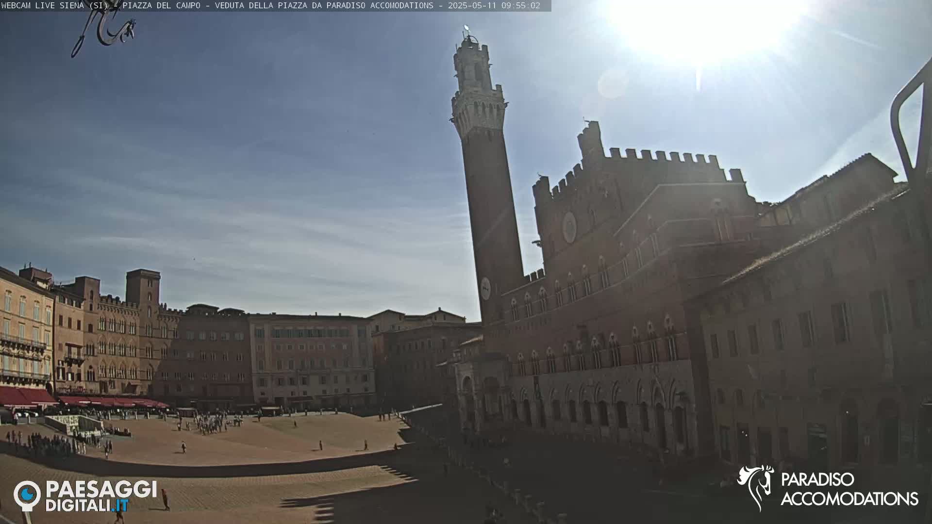 A sunny day in a large, brick-paved plaza surrounded by medieval-style buildings, including a tall tower.