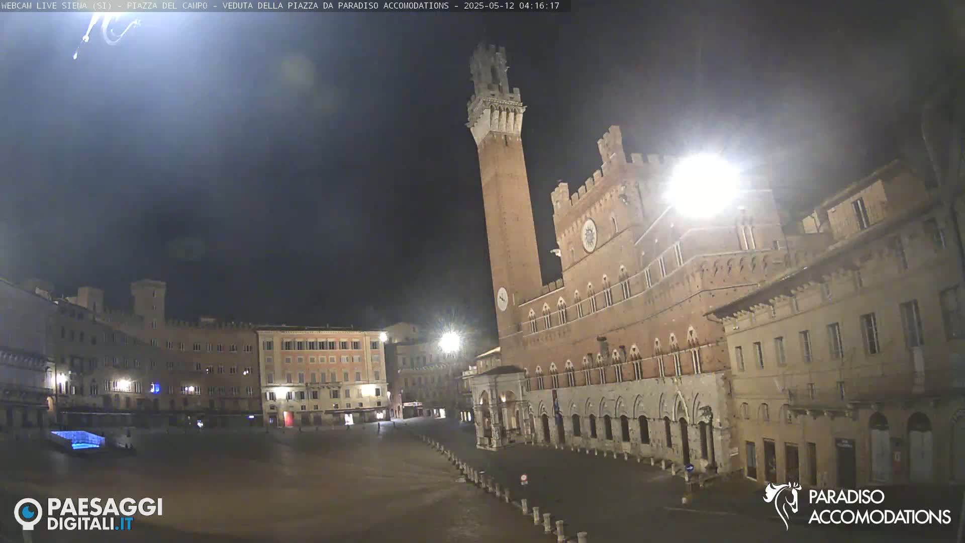 A large, empty plaza at night is surrounded by historical brick buildings, one prominently featuring a tall tower.