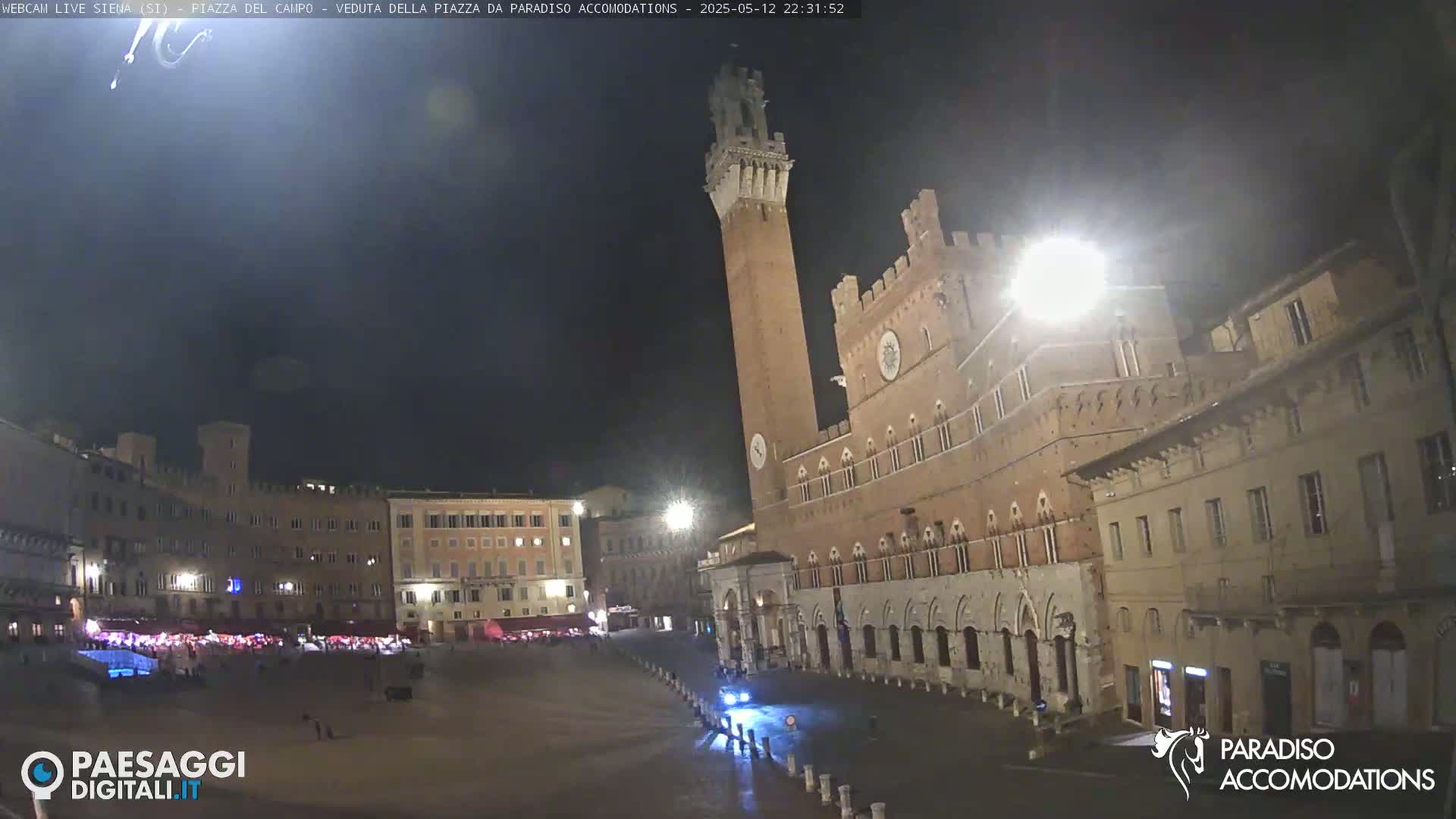 A large, empty plaza at night is surrounded by historic brick buildings, one of which features a tall tower.