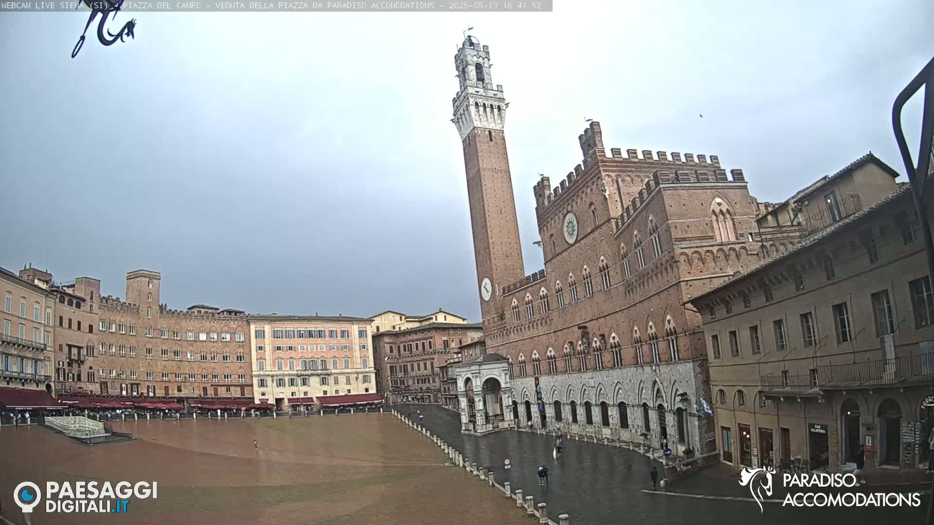 A large, brick public square in a town, possibly in Italy, is shown on an overcast day, with a tall tower and a large brick building in the background.