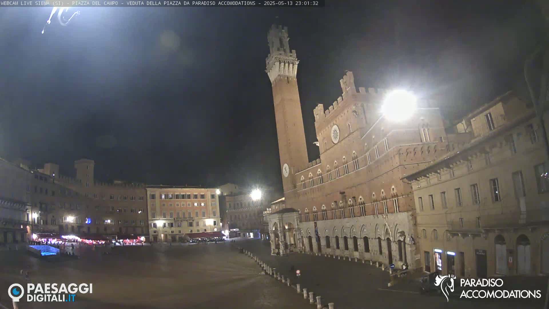 A large, empty plaza at night is surrounded by historic-looking buildings, including a tall brick tower.
