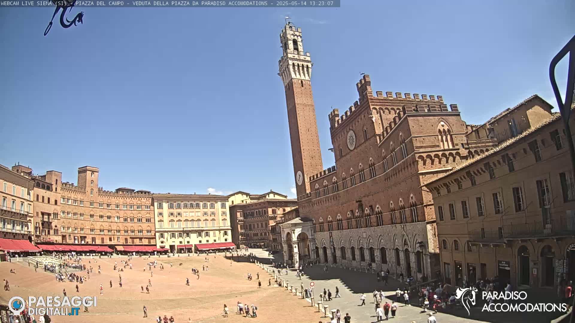 A large, sunny plaza is surrounded by historic brick buildings, including a tall tower and a palazzo, with people scattered throughout the square.