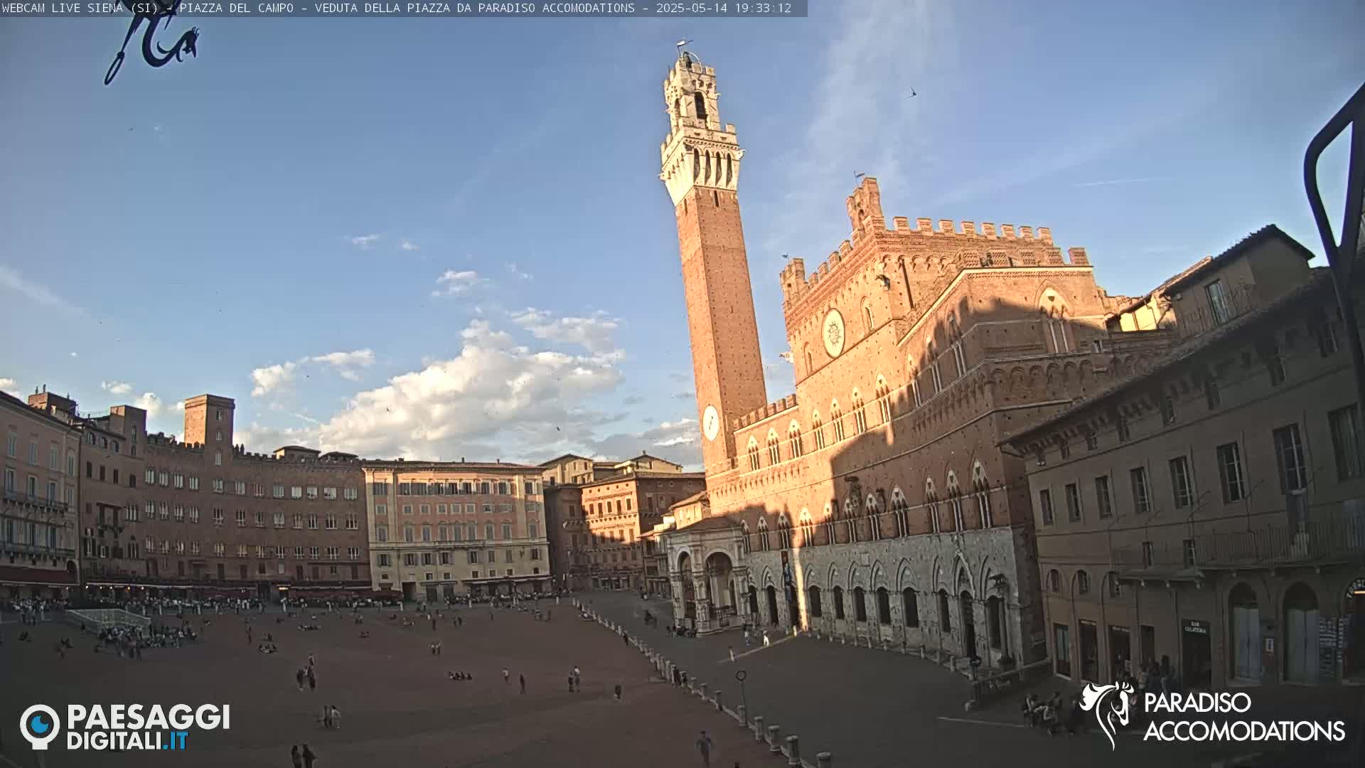 A large, brick piazza in Siena, Italy, is mostly empty under a partly cloudy, late afternoon sky; a tall tower and a large brick building are prominent features.