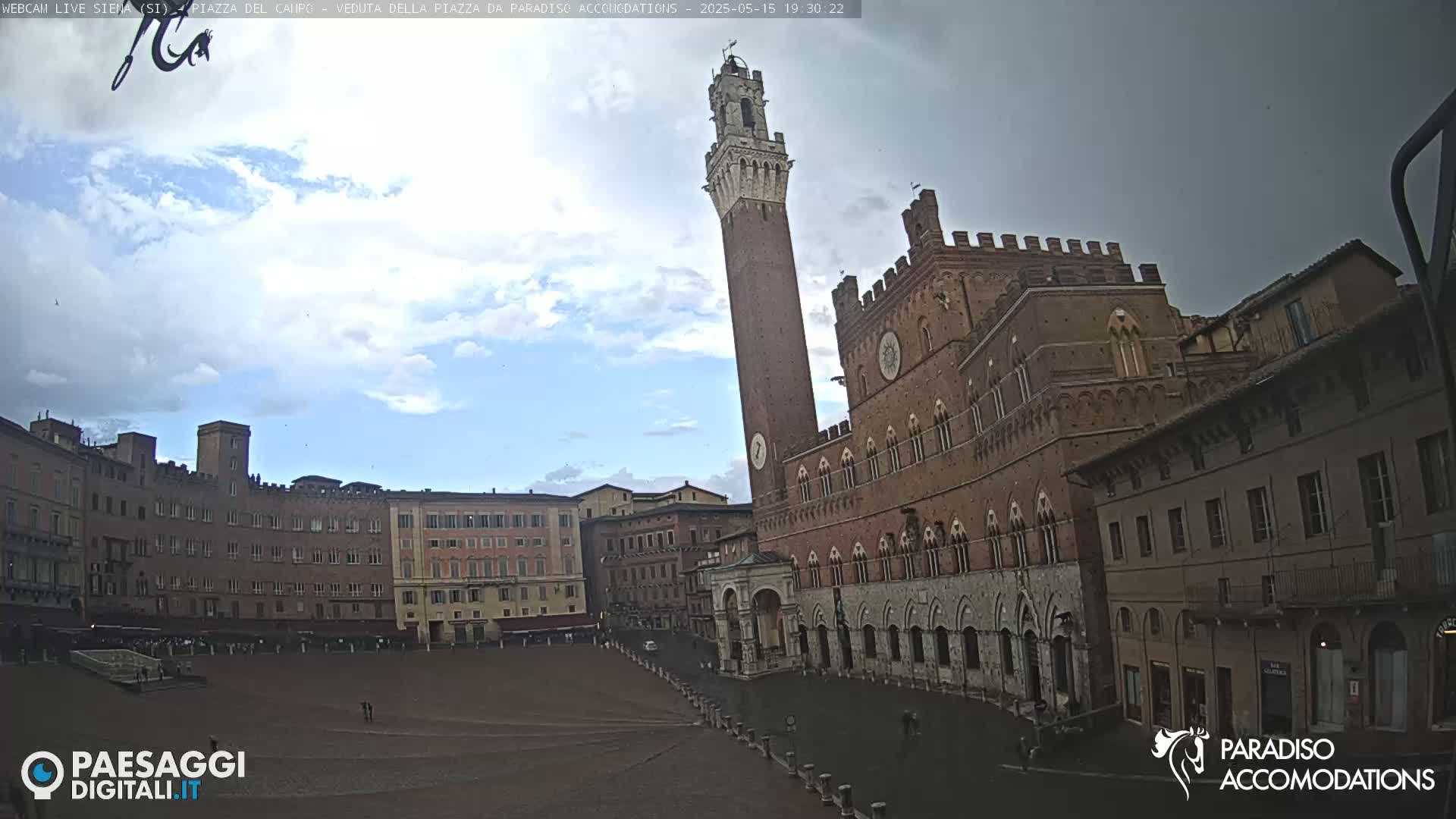 A mostly cloudy sky overlooks a large, empty piazza in Siena, Italy, surrounded by medieval buildings, including a tall brick tower.