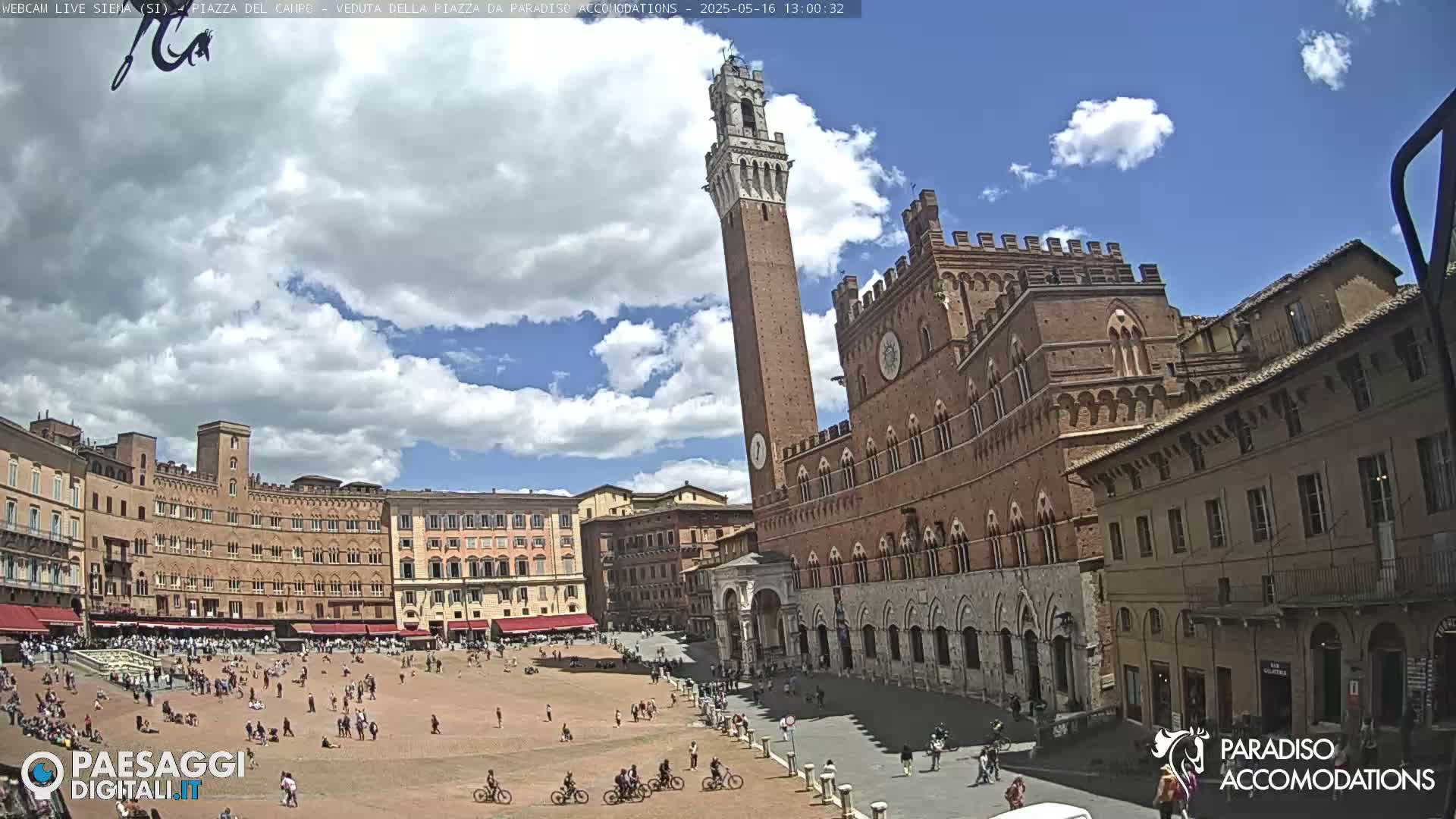 A mostly sunny day shows a large brick plaza with many people and bicycles, surrounded by historic buildings, including a tall, slender tower.