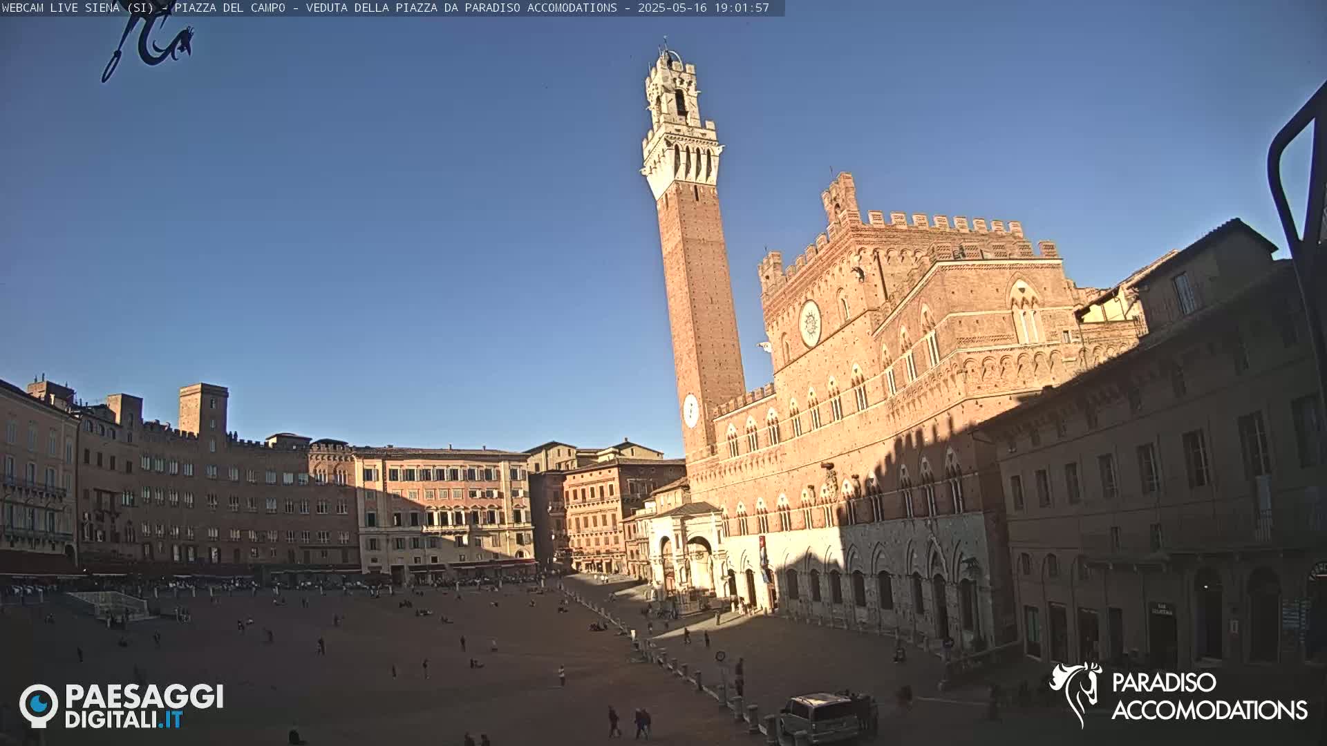 A large, sunlit piazza in Italy is surrounded by medieval brick buildings, including a tall tower and a grand palace.