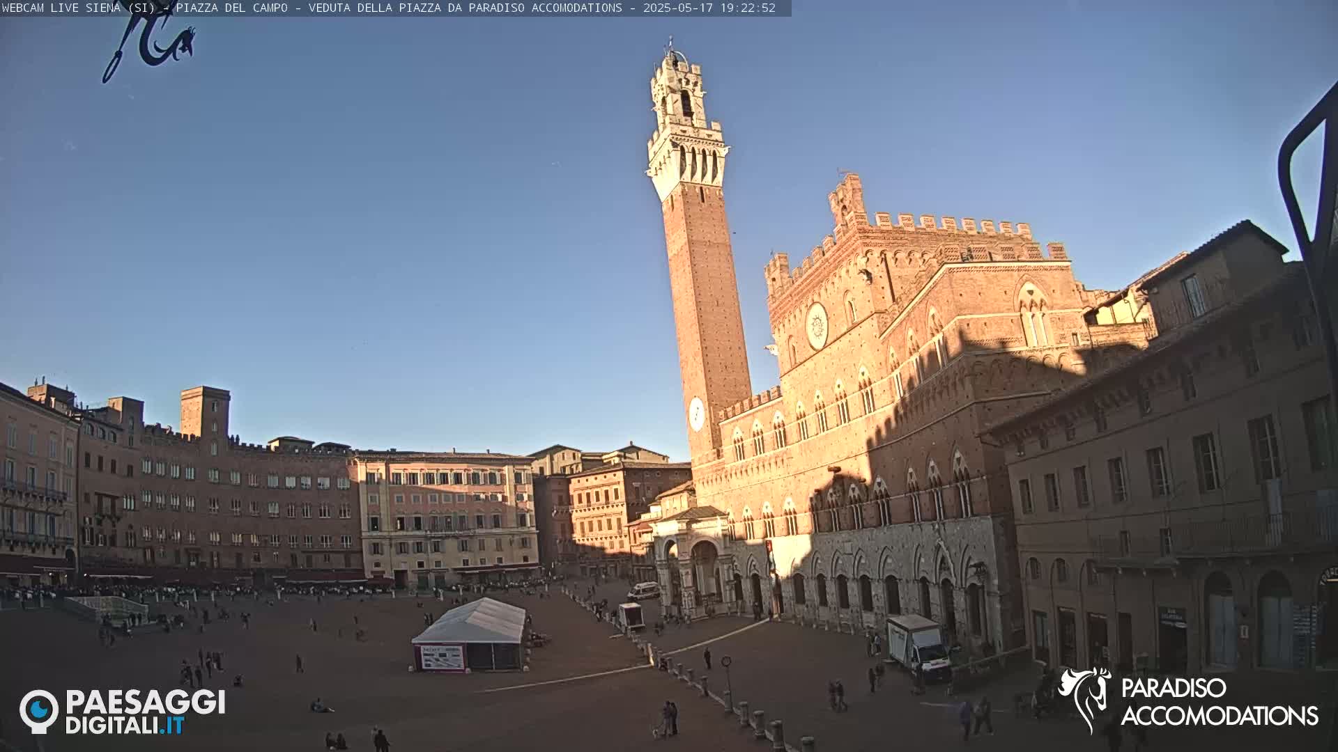 A large, brick public square in Siena, Italy, is mostly empty under a clear, late-afternoon sky, with a tall tower and surrounding buildings visible.