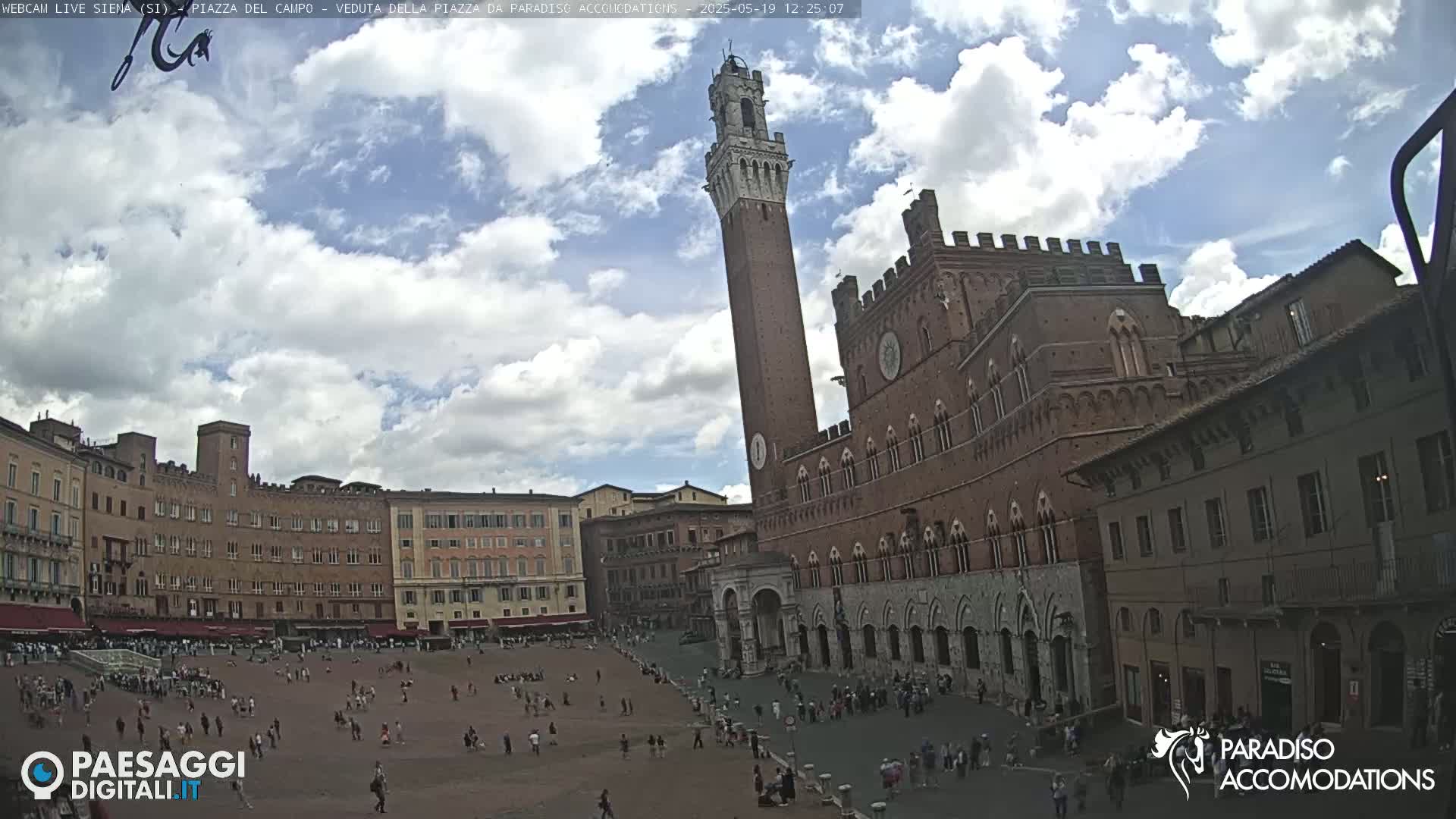 A large, brick public square with a tall tower and several historical buildings is visible under a partly cloudy sky.