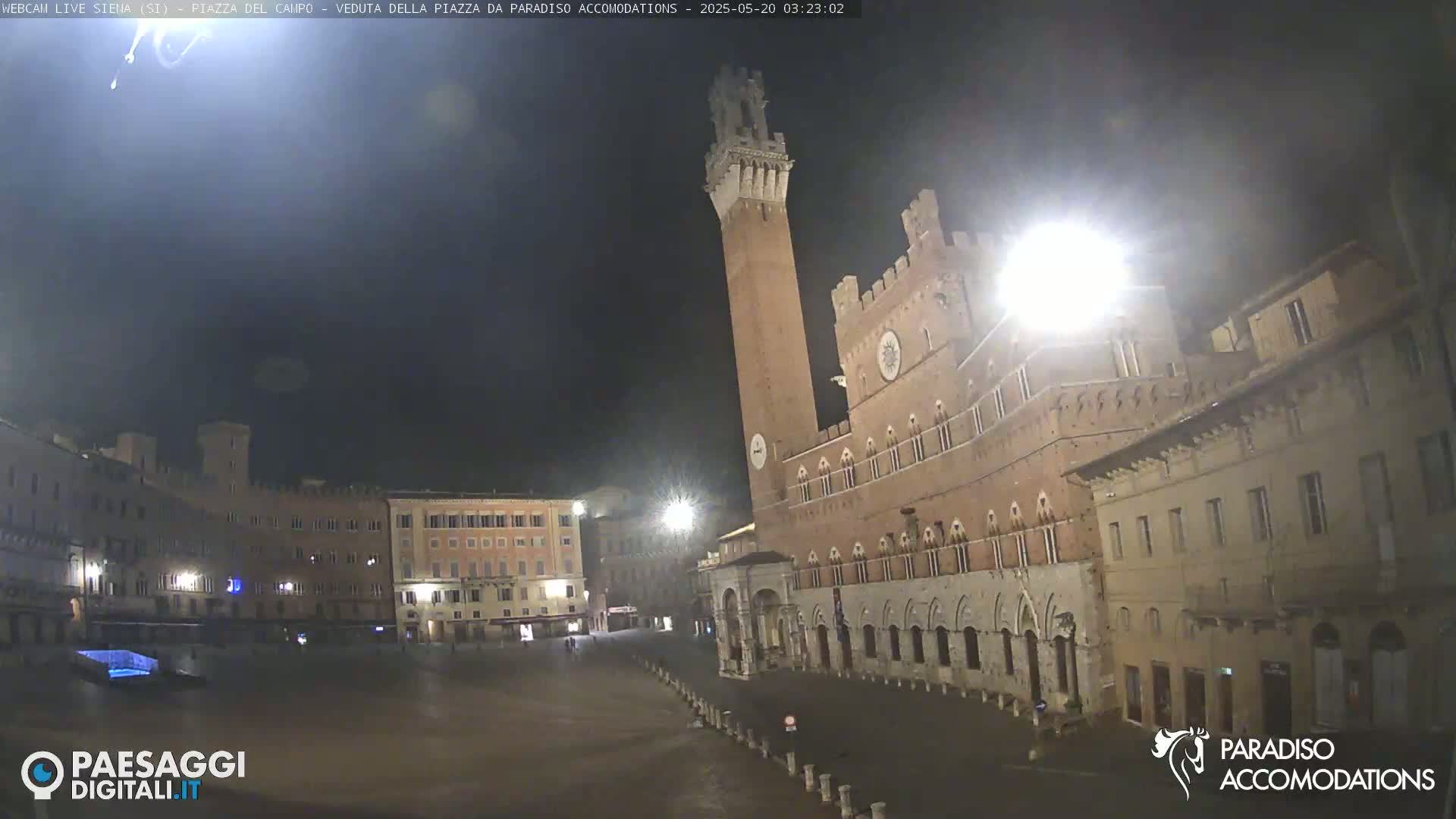 A mostly empty, large square at night, surrounded by old, illuminated buildings, including a tall tower and a large palace.