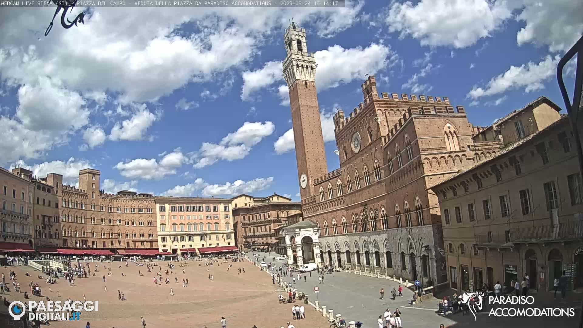 A large, mostly empty piazza in a sunny city is surrounded by medieval-style buildings, including a tall, slender tower.