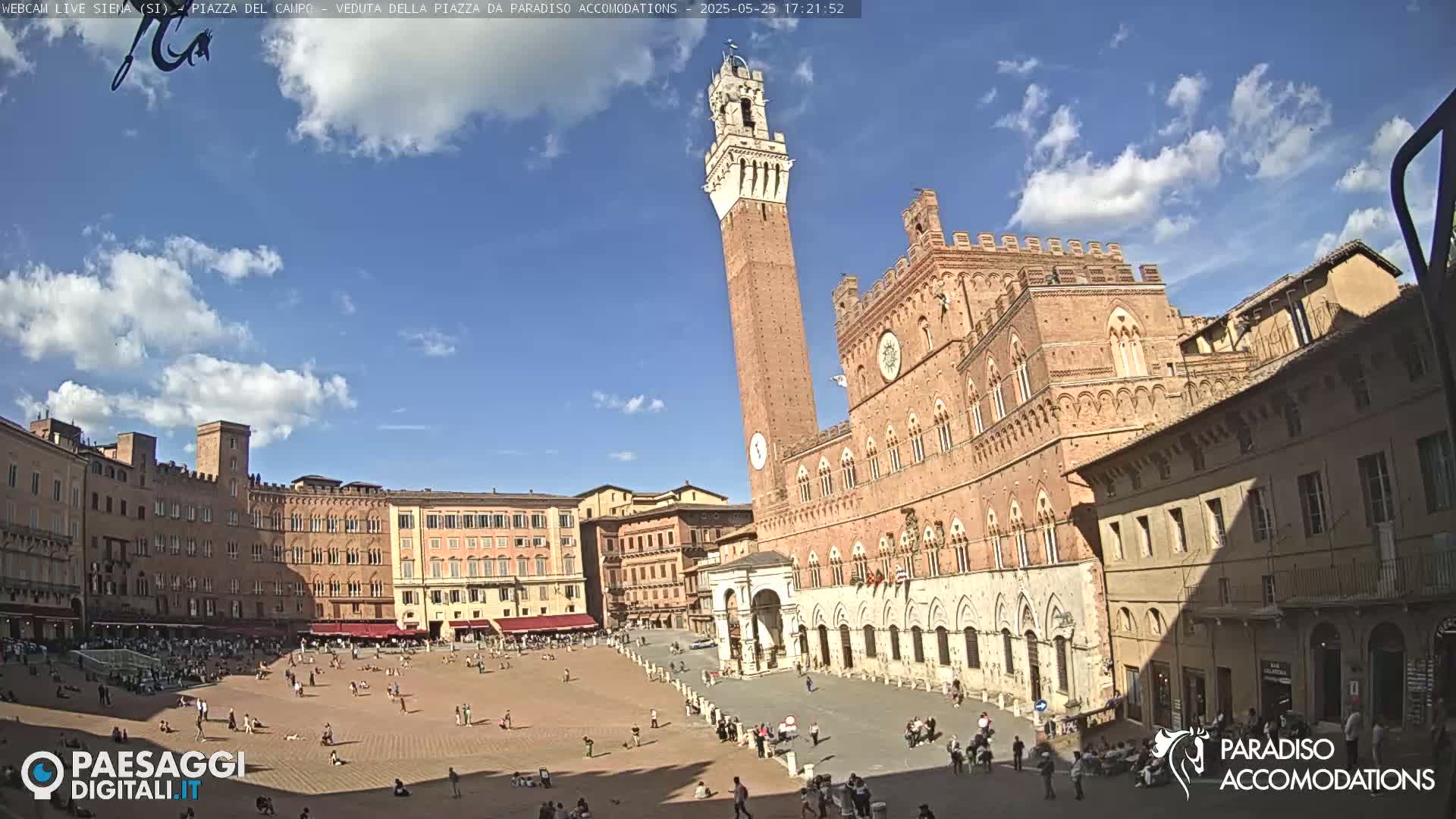A large, brick piazza in Siena, Italy, is filled with people under a mostly sunny sky with a few clouds.