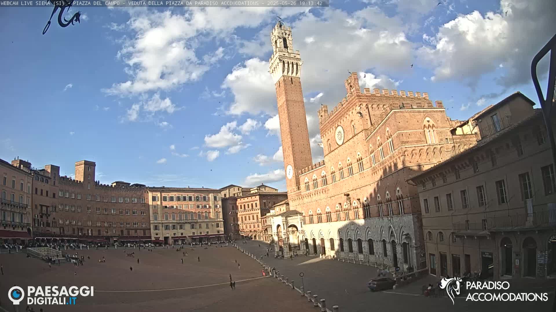 A large, brick public square is shown under a partly cloudy sky, with a tall, slender tower and a large, ornate building prominent in the scene.