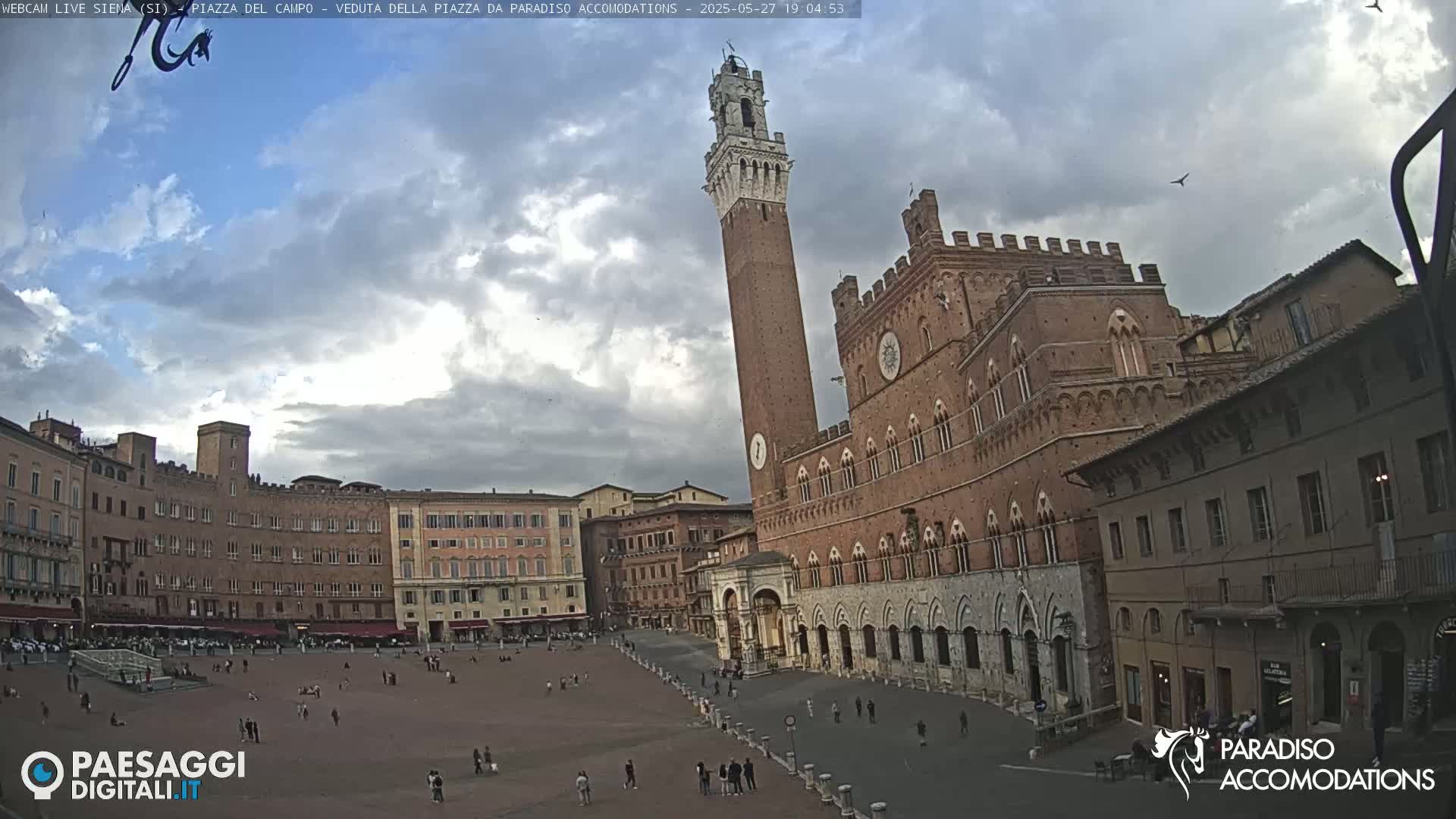 A large, open square in a historic European city is surrounded by brick buildings, including a tall tower and a large palace, under a partly cloudy sky.