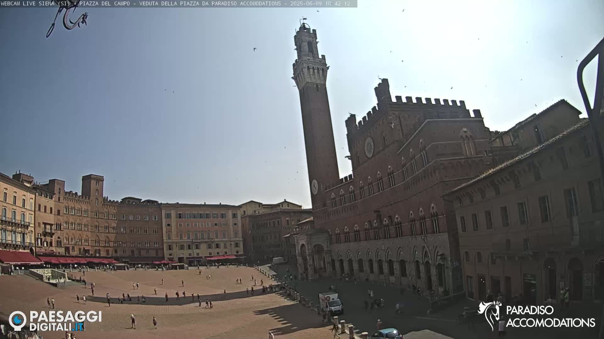 A large, mostly empty plaza is surrounded by medieval-style buildings under a clear, sunny sky.
