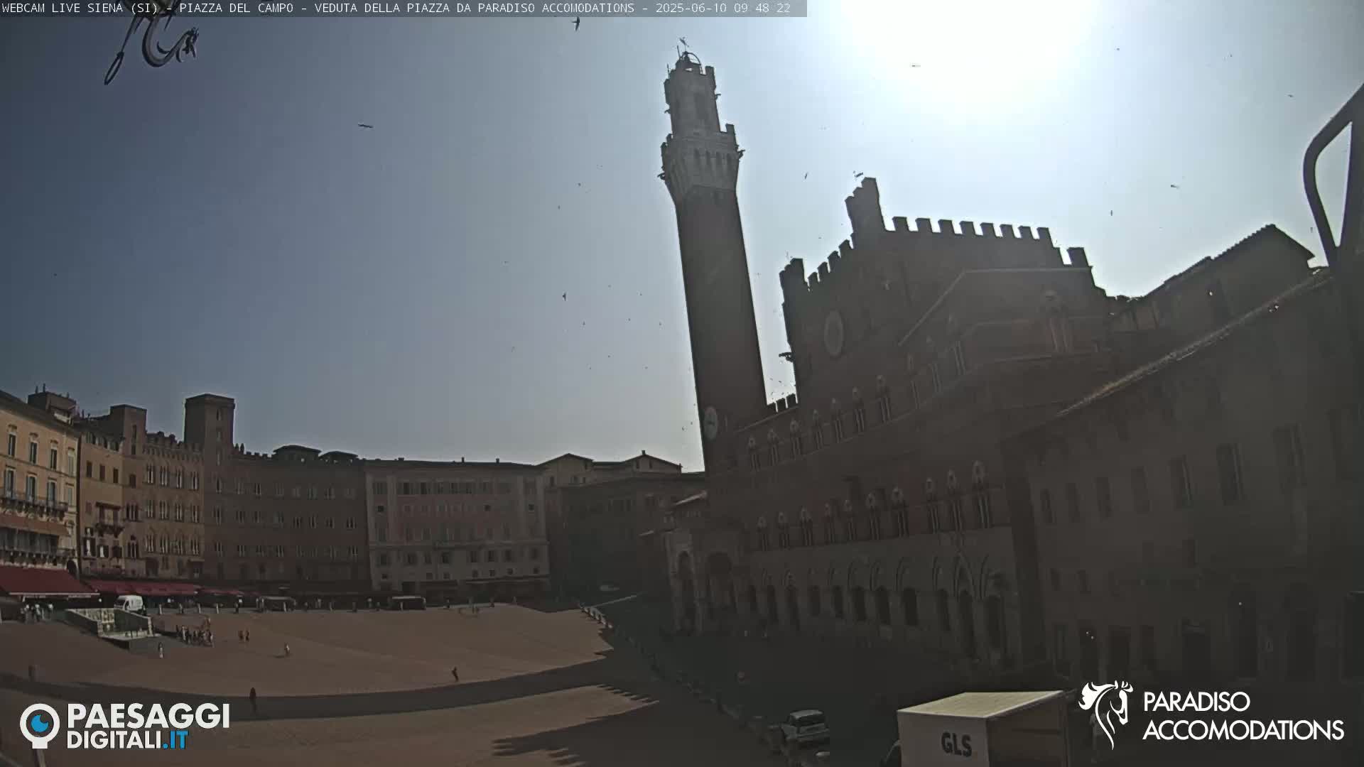 A sunny day in a large, empty plaza surrounded by medieval-style buildings, one of which features a tall tower.