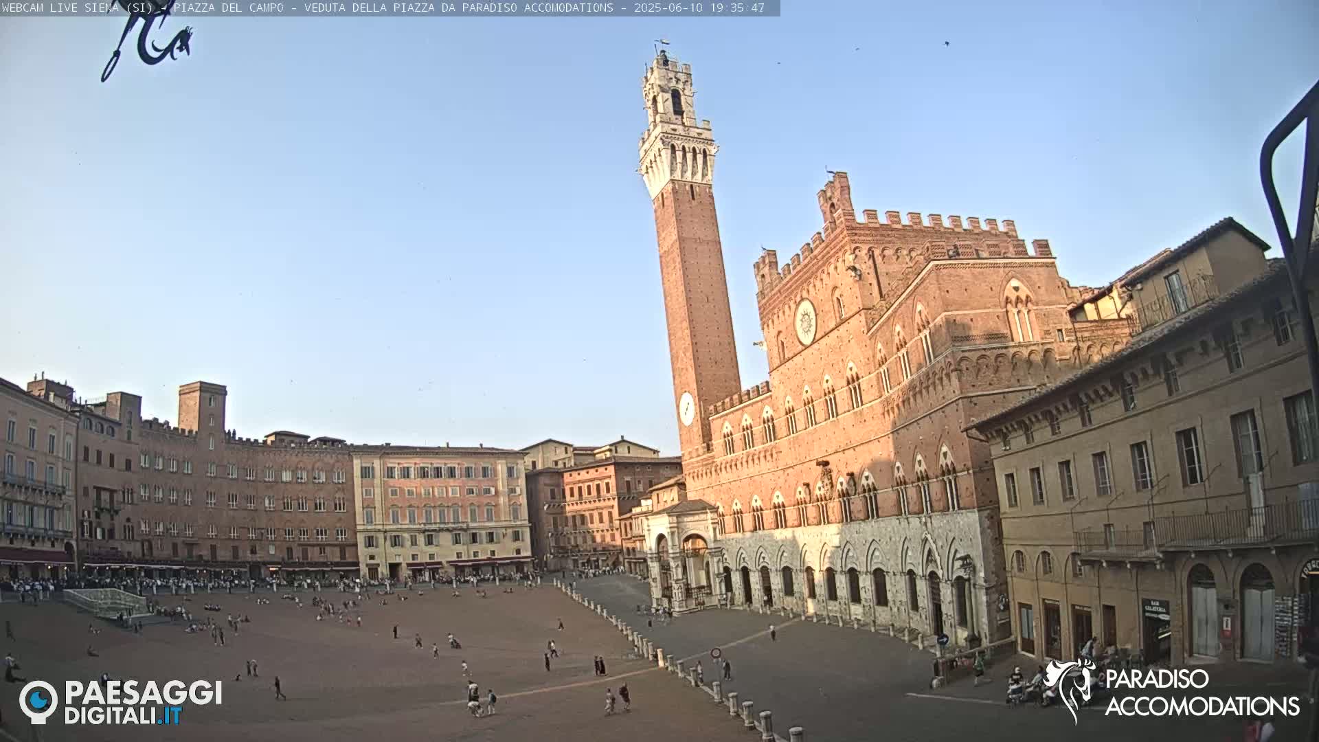 A large, mostly empty plaza is surrounded by historic brick buildings, including a tall tower and a grand palazzo, under a clear, bright sky.