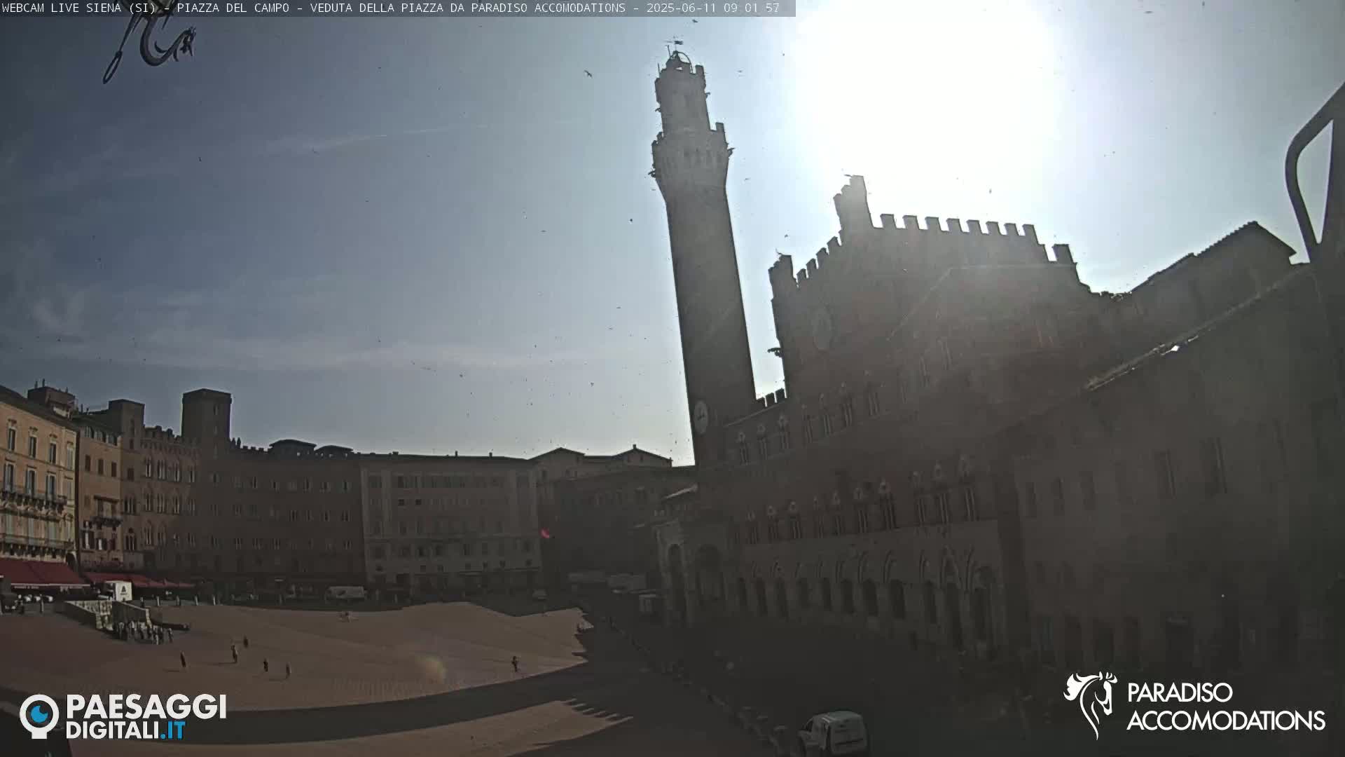 A sunny, mostly clear day reveals a large, mostly empty plaza surrounded by historical-looking buildings, including a tall tower.