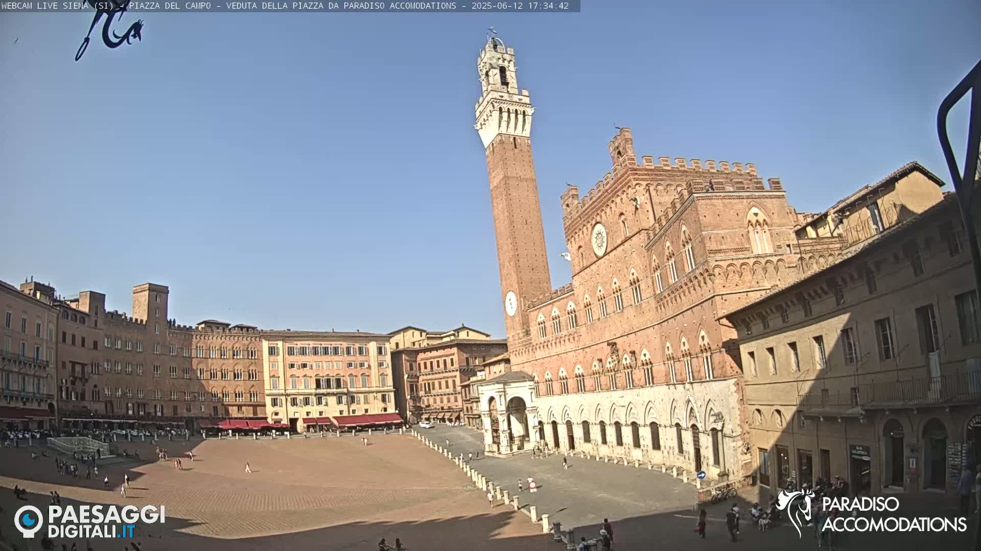 A large, brick piazza with a tall tower and surrounding medieval buildings under a clear, sunny sky.