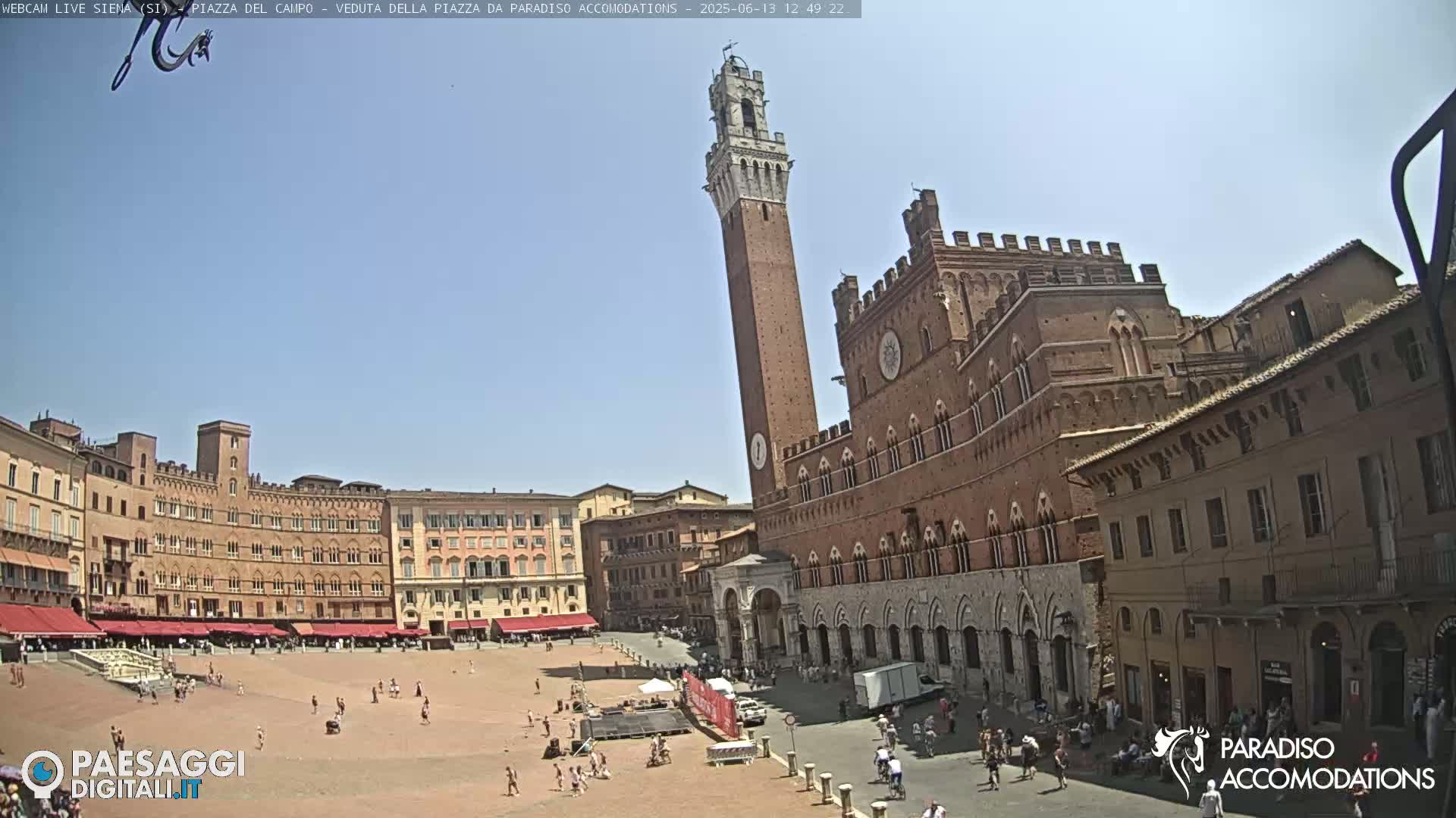 A large, sunny plaza in Italy is surrounded by historic brick buildings, one featuring a tall tower.