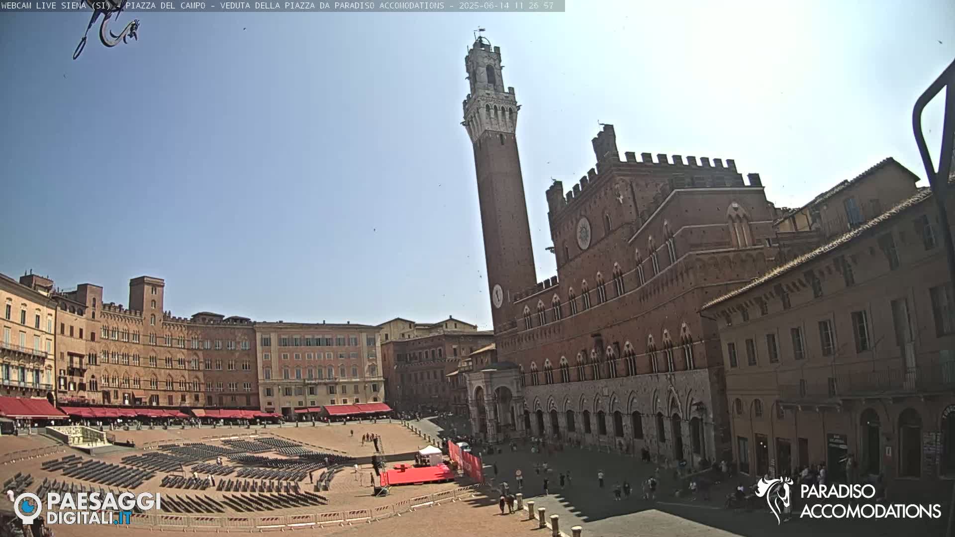 A large, brick public square in a city, under a clear blue sky, features a tall tower and several medieval-style buildings, with rows of empty chairs set up in the square.