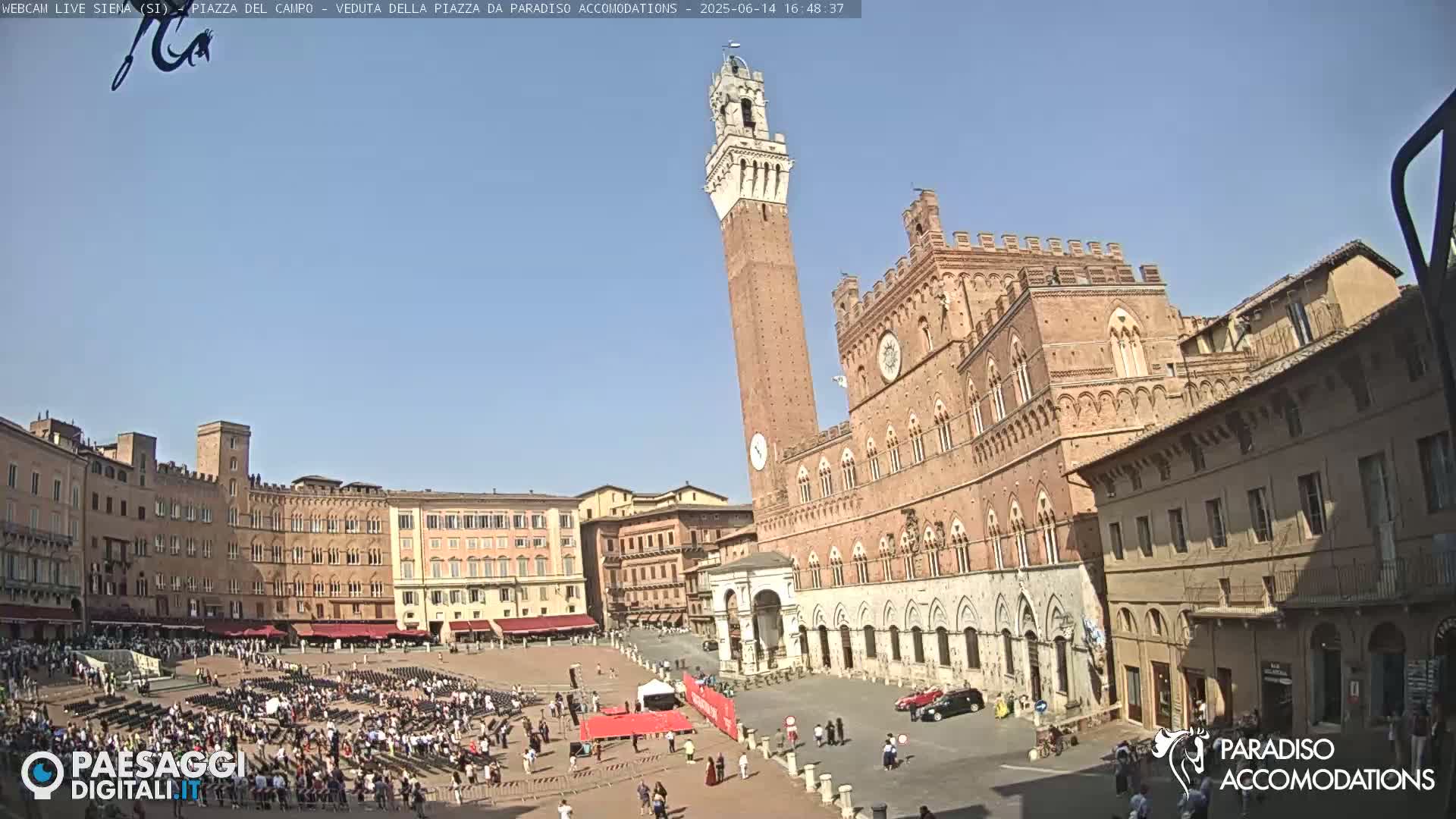 A large, mostly empty town square is surrounded by historic brick buildings under a clear, sunny sky, with a tall tower prominently featured in the center.