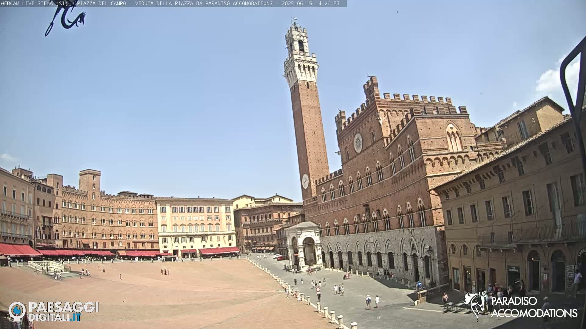 A large, brick piazza in bright sunlight, featuring a tall tower and several historical buildings surrounding the open space with a few people scattered about.