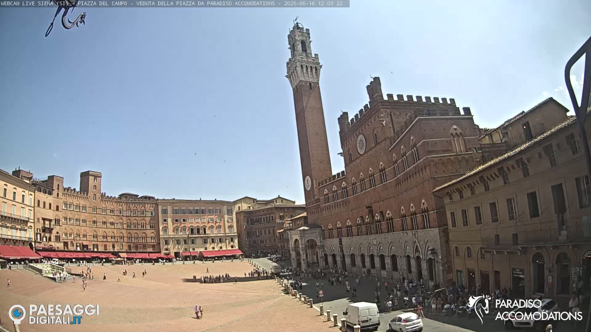 A large, brick piazza in Siena, Italy, is shown on a sunny day, with a tall tower and surrounding buildings visible.