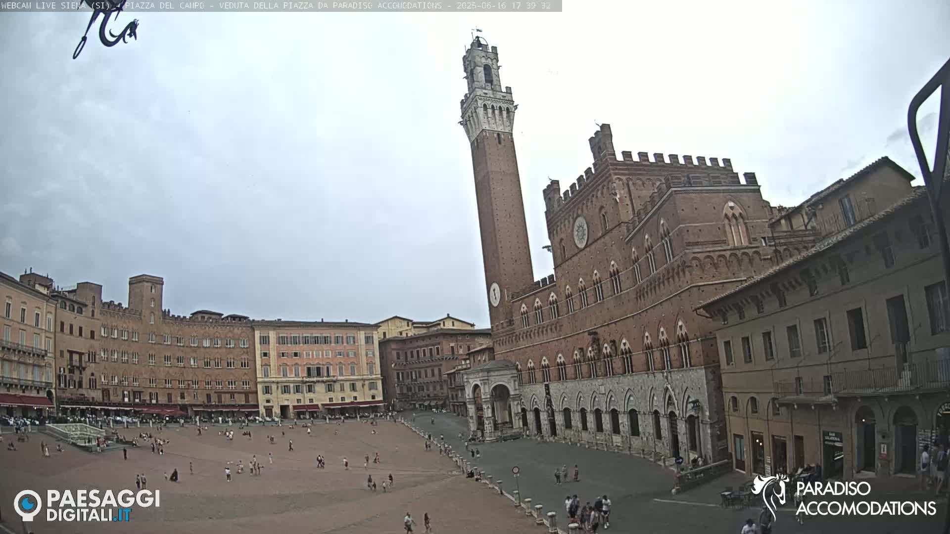A large, mostly empty plaza in a city with several brick buildings, including a tall, slender tower, under a cloudy sky.