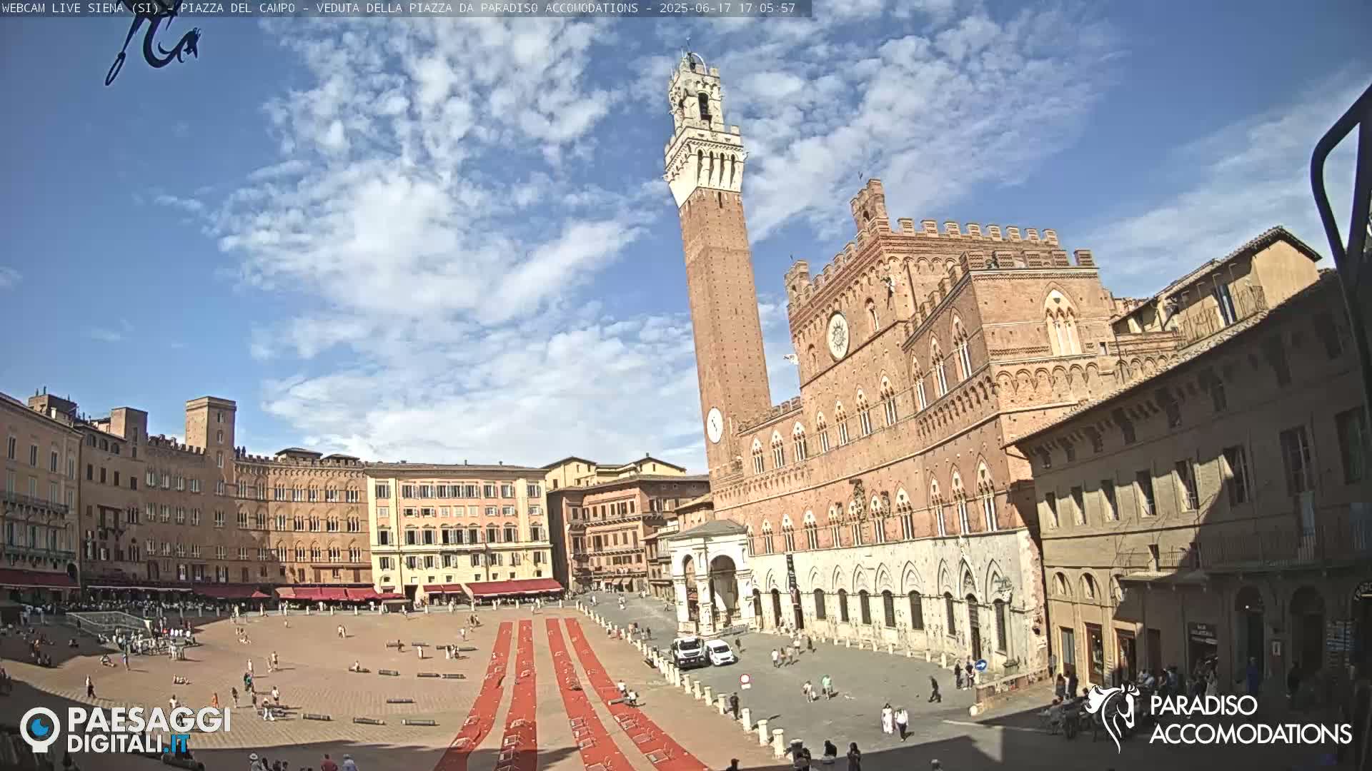 A large, sunny town square is filled with people and red carpeting, surrounded by tall, terracotta-colored buildings, including a prominent tower.