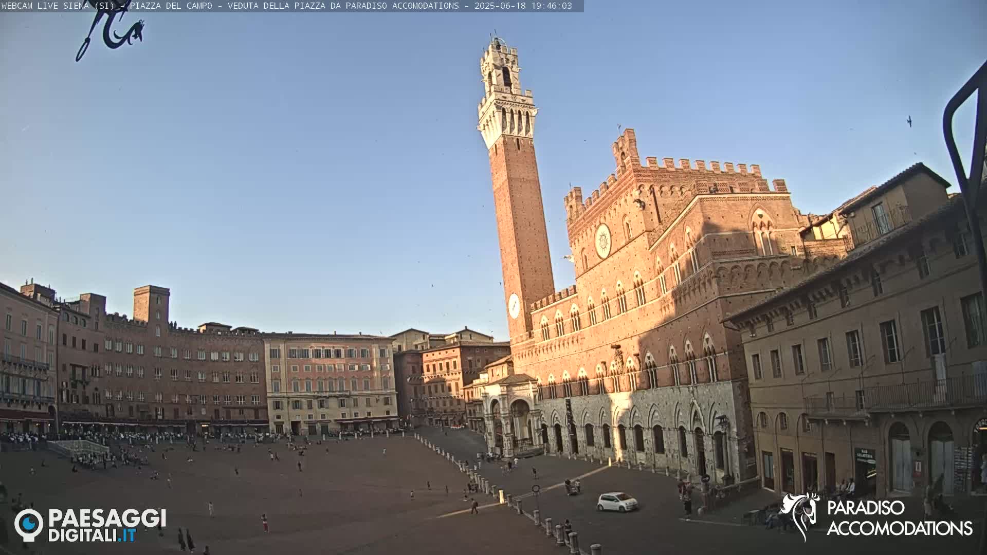 A large, brick piazza in Siena, Italy, is filled with people under a clear, late afternoon sky, with the Torre del Mangia and Palazzo Pubblico prominently featured.