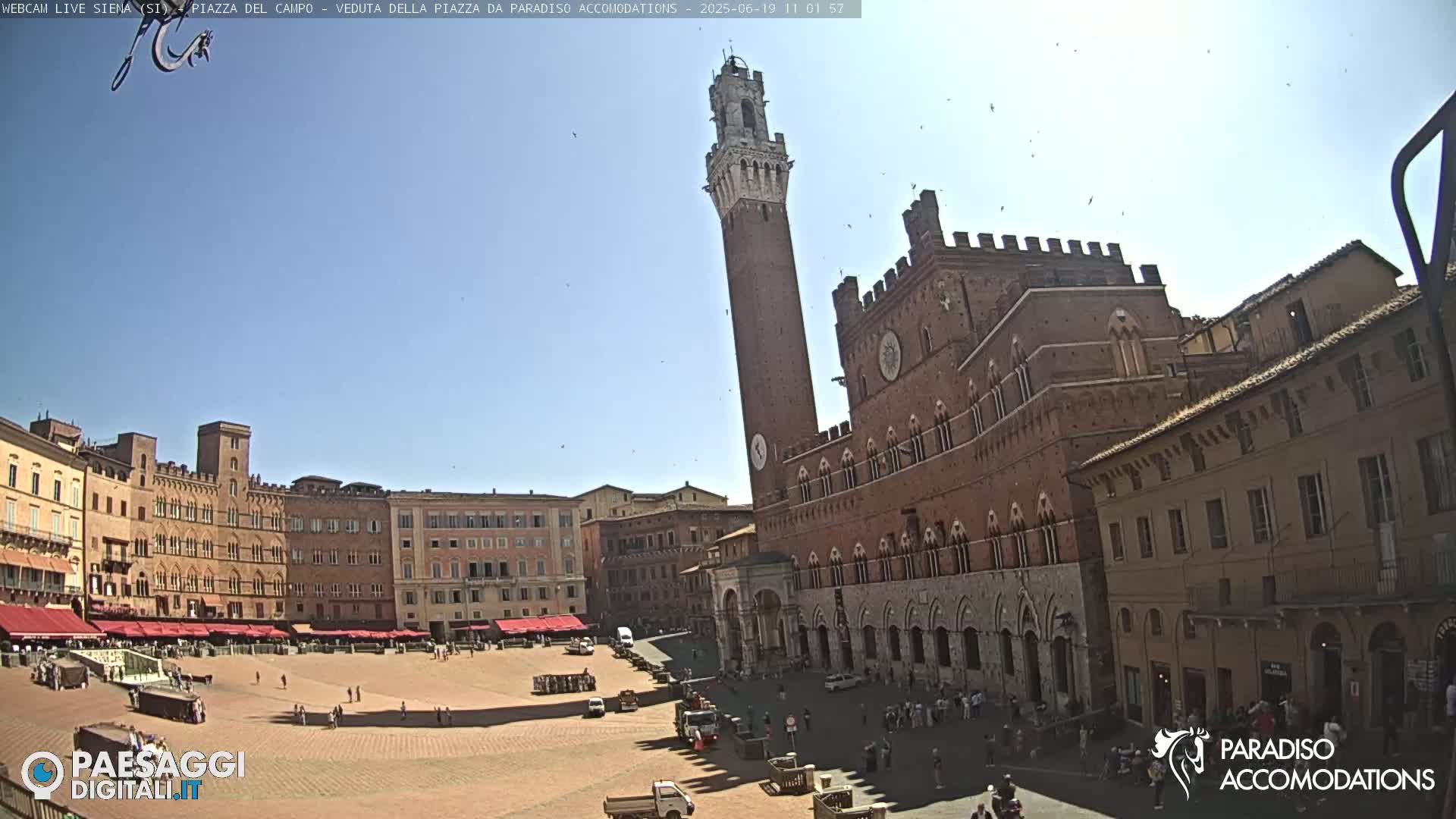 A large, sunlit piazza in Italy, surrounded by historic brick buildings, including a tall tower and a palace, with a few people and vehicles visible.