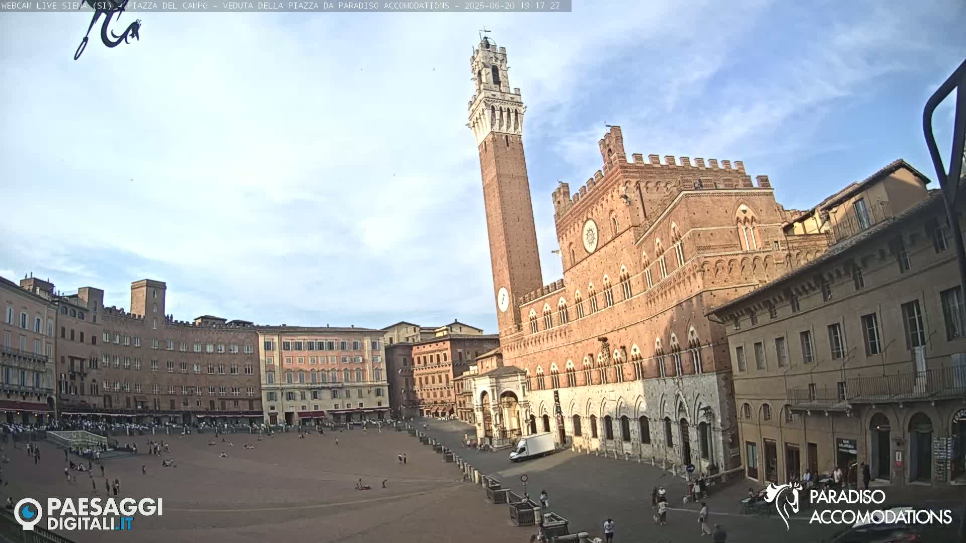A large, mostly empty town square is surrounded by historic brick buildings, including a tall tower, under a partly cloudy sky.