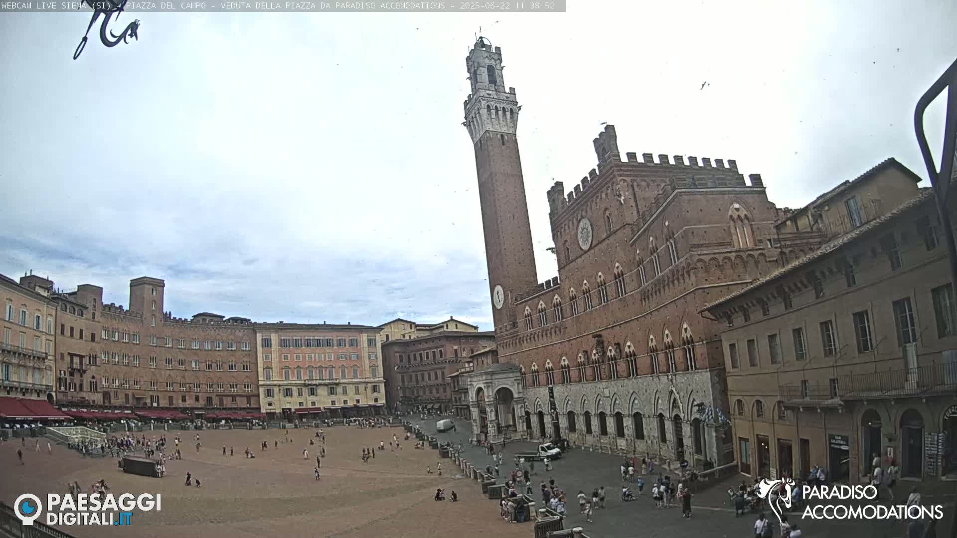 A large, mostly empty plaza is surrounded by historic brick buildings, including a tall tower, under a mostly cloudy sky.