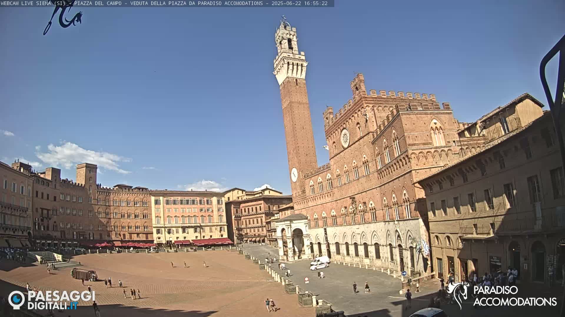 A large, brick public square is seen on a sunny day, with several historical buildings surrounding it, including a tall tower.