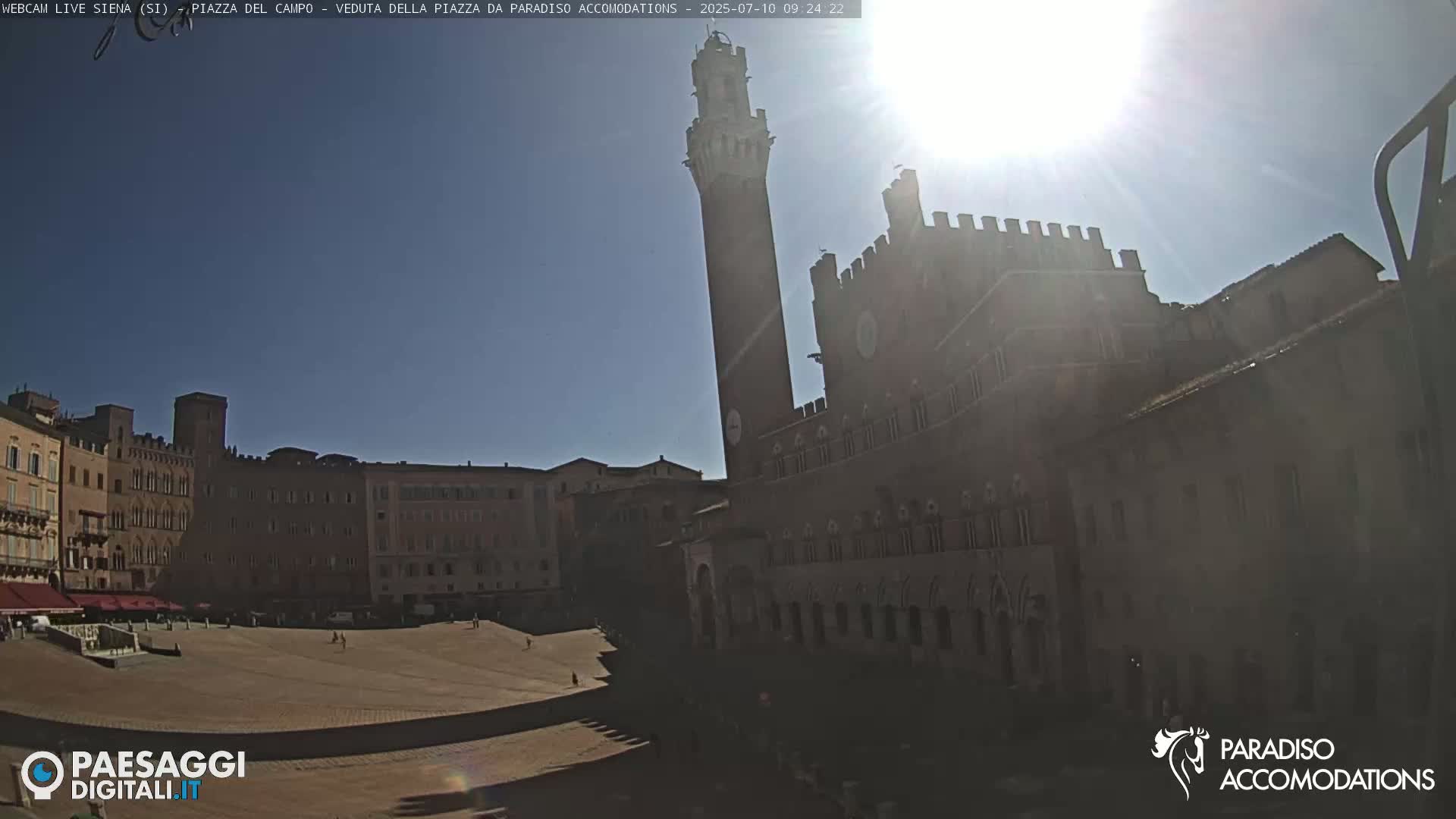A large, empty plaza is surrounded by historic brick buildings under a bright, sunny sky.
