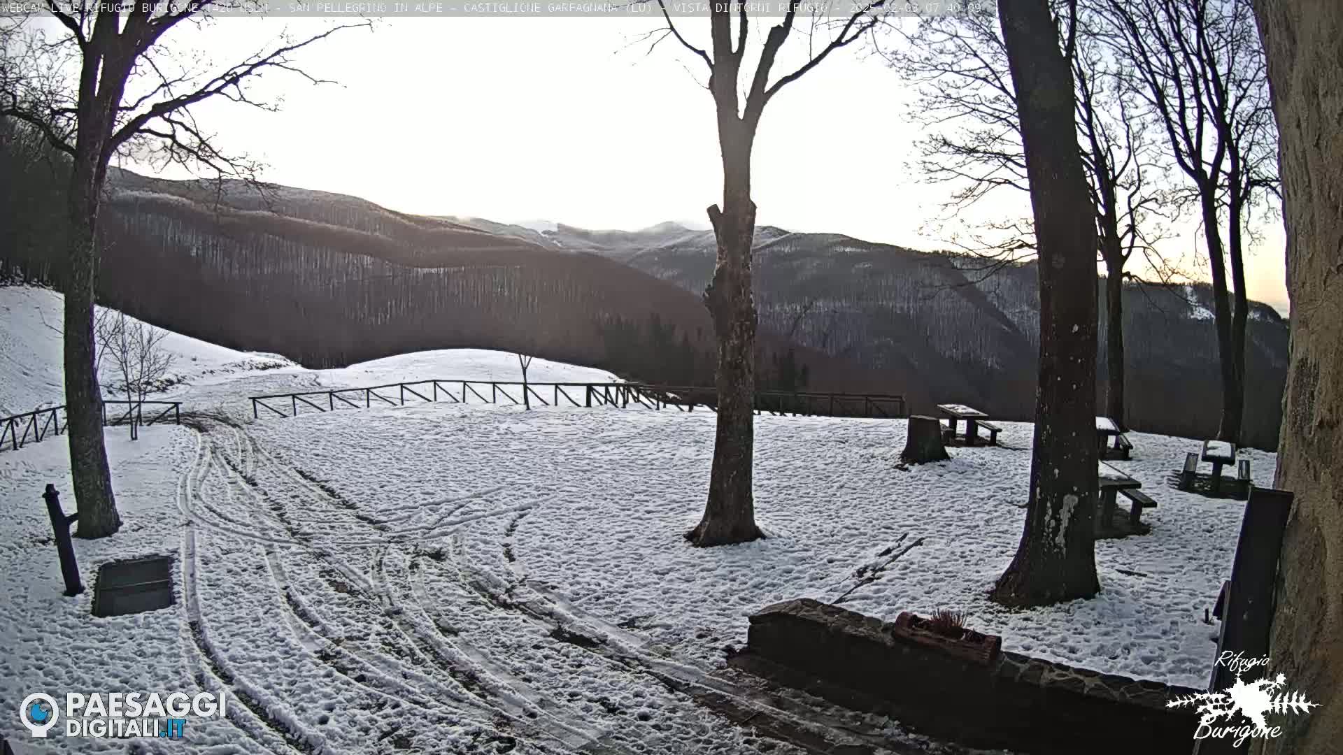 A snow-covered area with several picnic tables surrounded by leafless trees and a view of distant, snow-dusted mountains under a twilight sky.