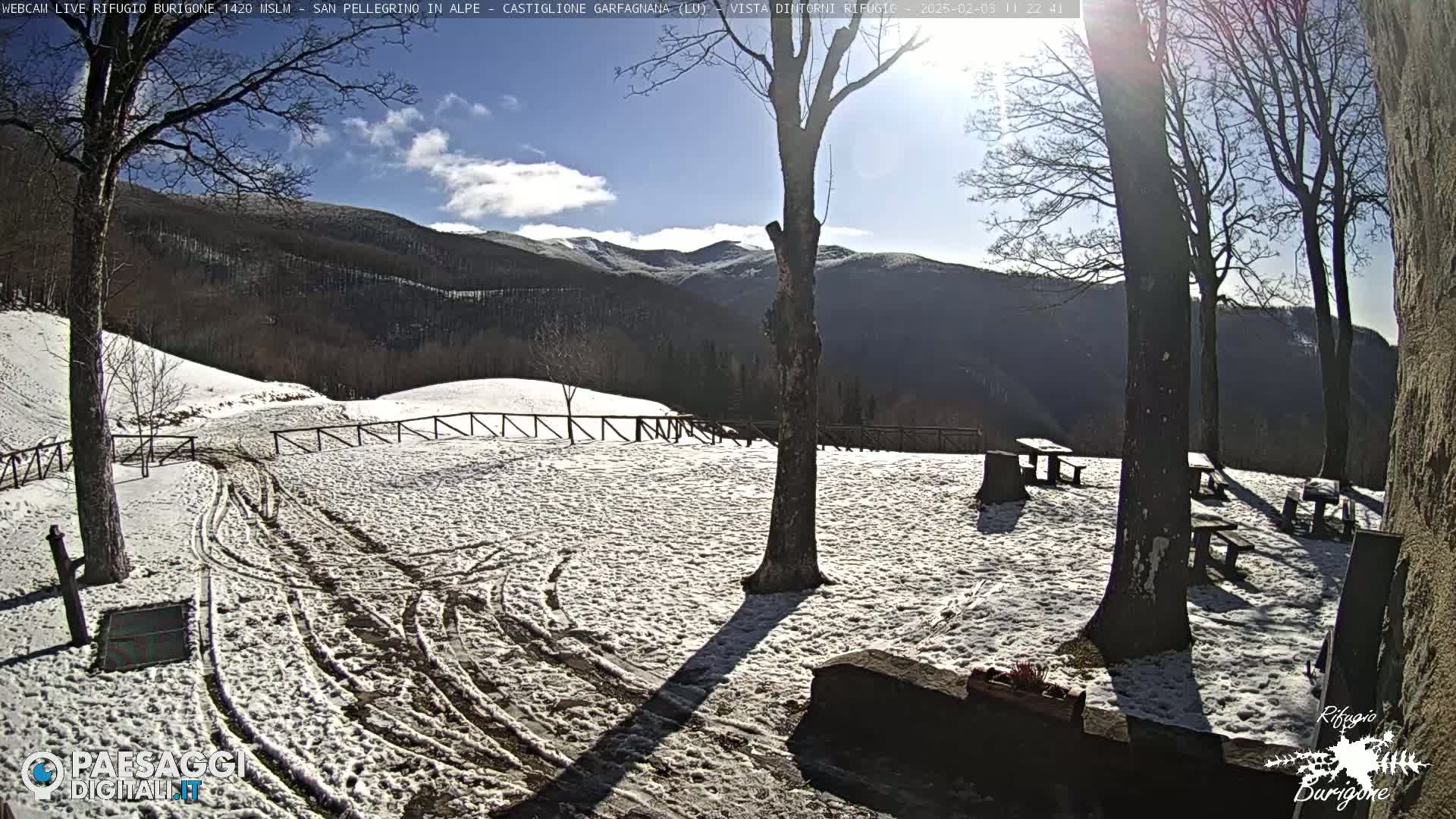 A snow-covered area with several picnic tables and trees, overlooking a mountain range under a sunny sky with some clouds.