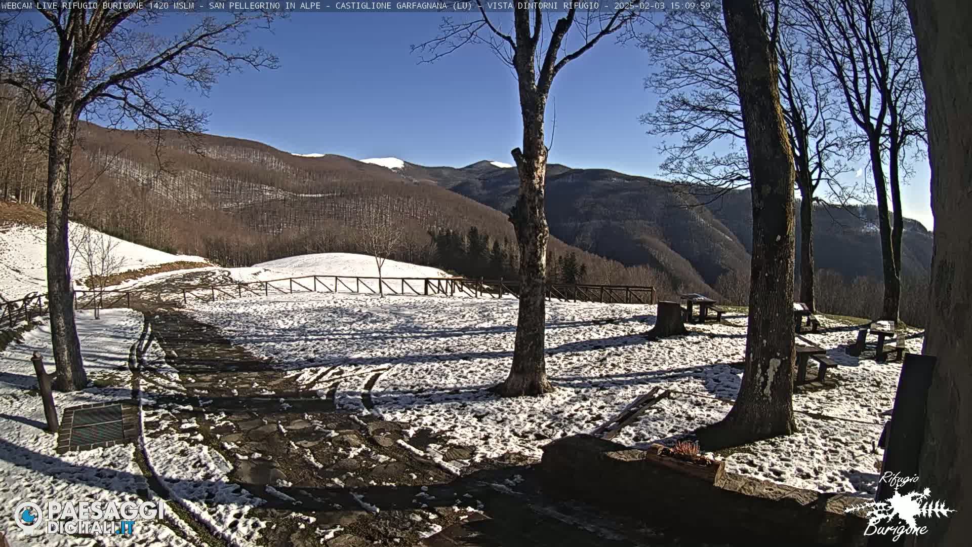 A snow-dusted, sunny mountainside scene with several picnic tables situated amongst bare trees, a partially snow-covered stone path, and a wooden fence.