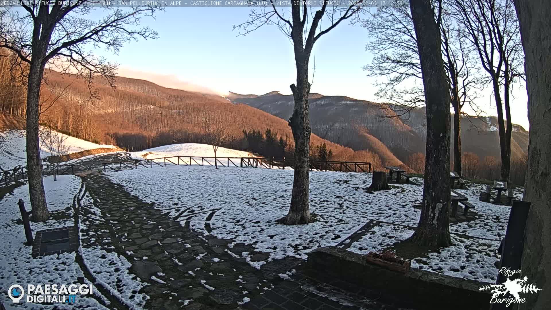 A snow-covered stone patio with several wooden picnic tables sits in front of a mountain vista under a clear, bright sky.