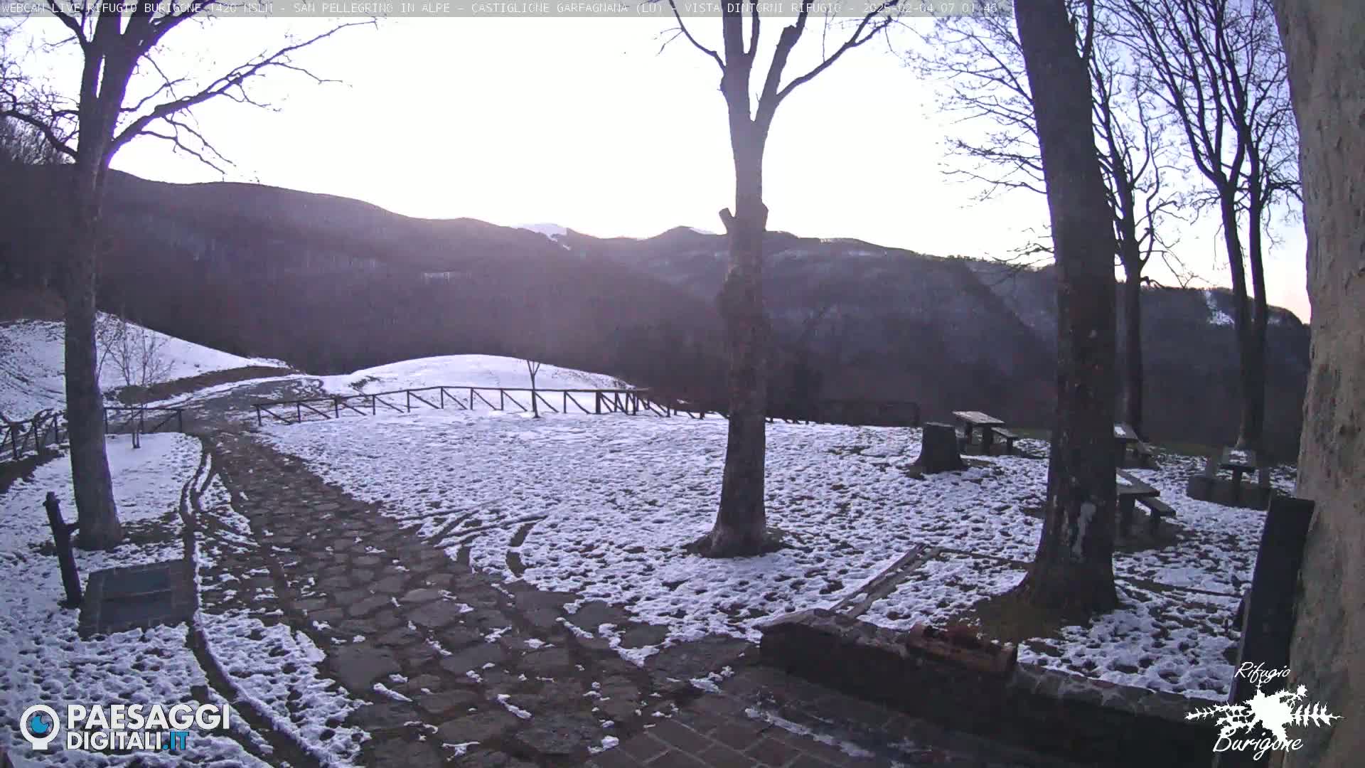 A snow-dusted stone path and picnic area are situated in front of a hilly, wooded landscape under a pale sky.