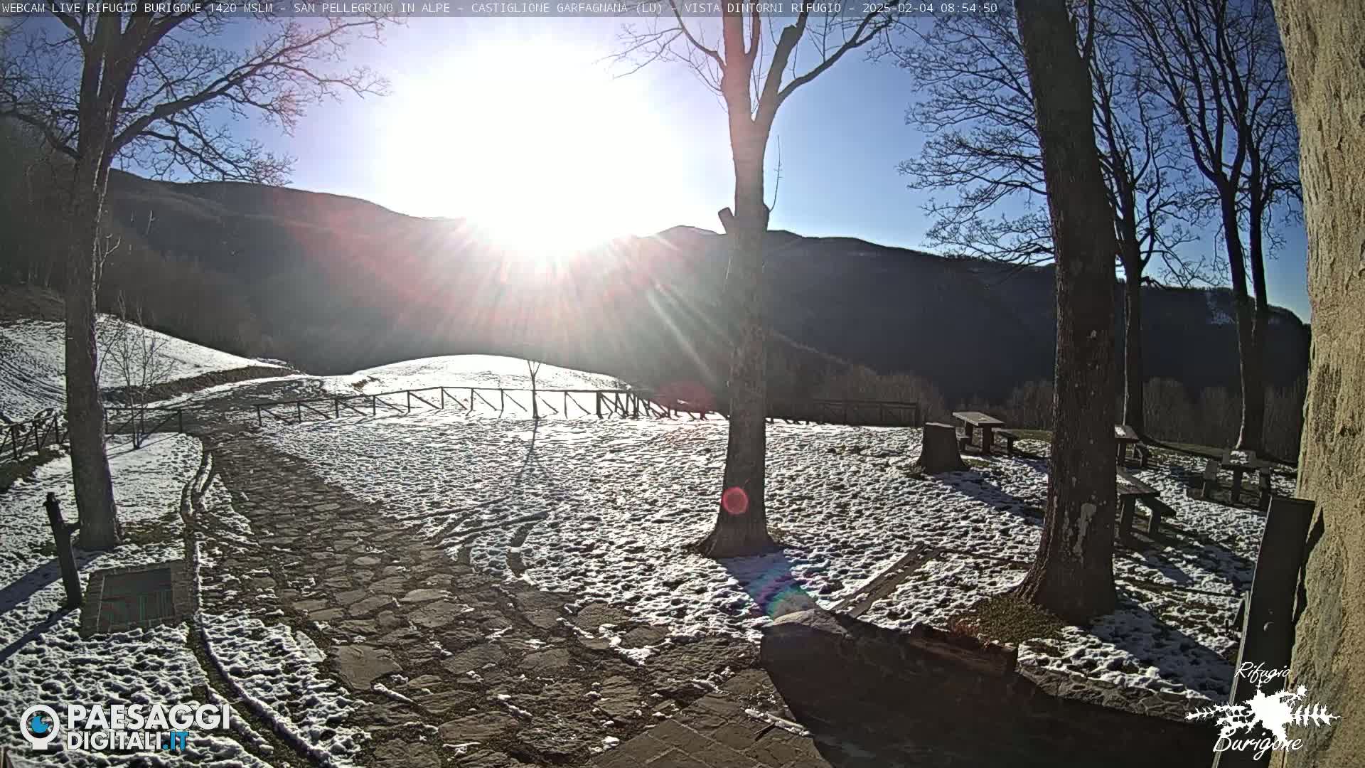 A snow-covered stone path and picnic area are situated beneath a bright sun in a mountainous region, with several bare trees casting shadows on the ground.