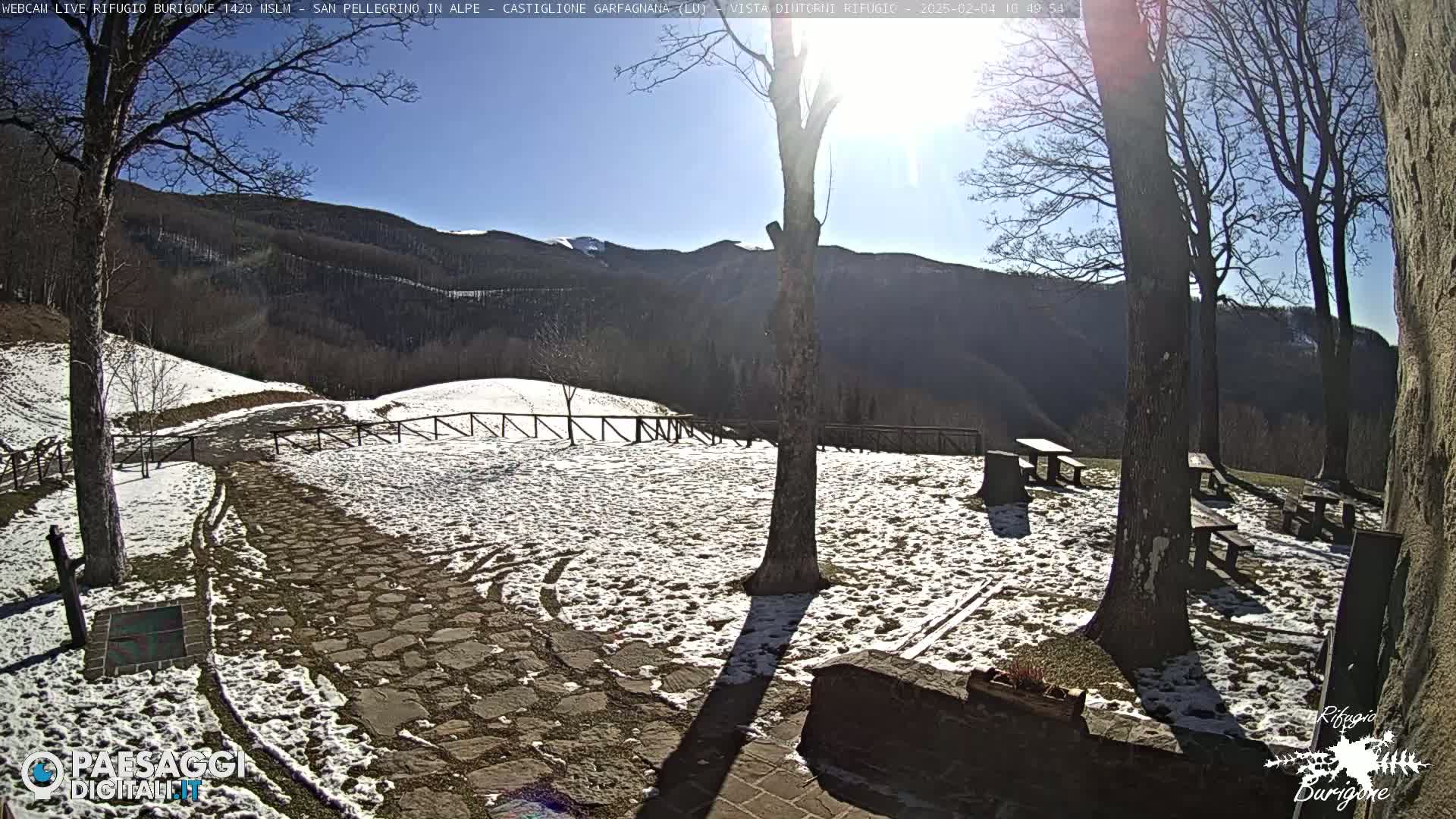 A sunny, snow-dusted stone patio area with several picnic tables sits below a mountain range under a clear blue sky.