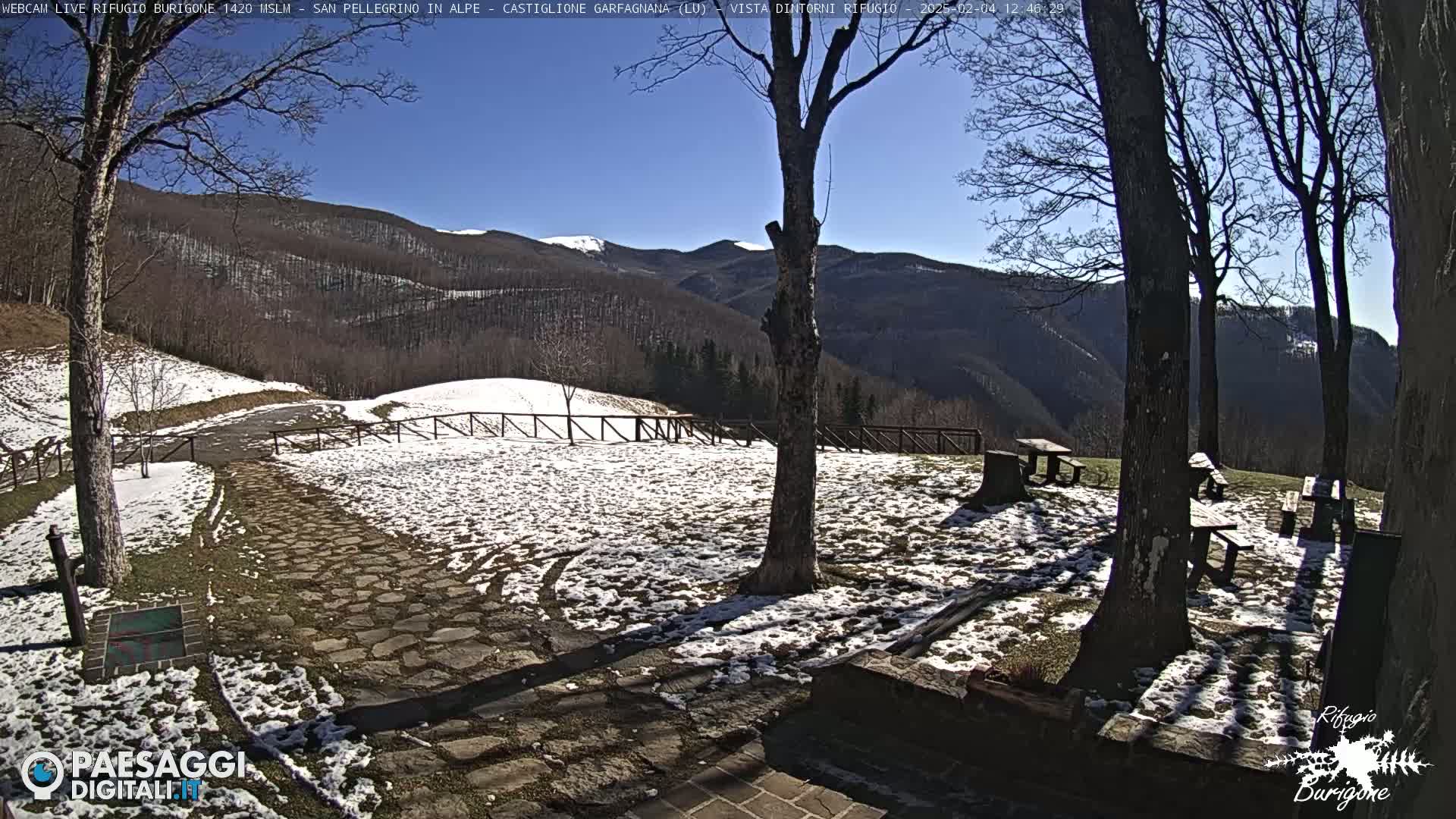 A stone pathway leads to a snow-dusted clearing with picnic tables nestled among bare trees, overlooking a mountain range under a bright, sunny sky.