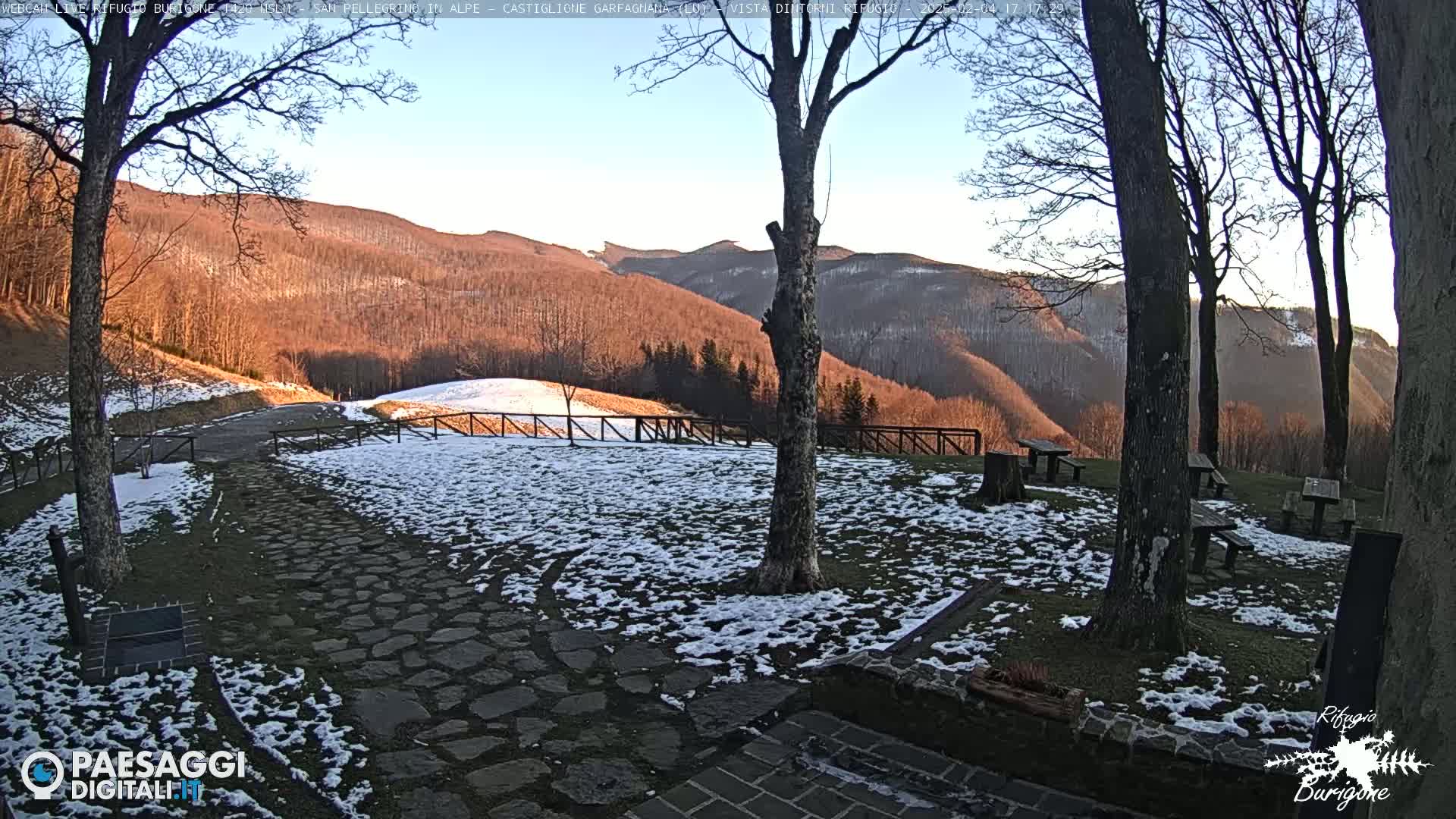 A snow-dusted stone path and grassy area with several picnic tables sit before a mountain range bathed in the warm light of late afternoon under a clear sky.